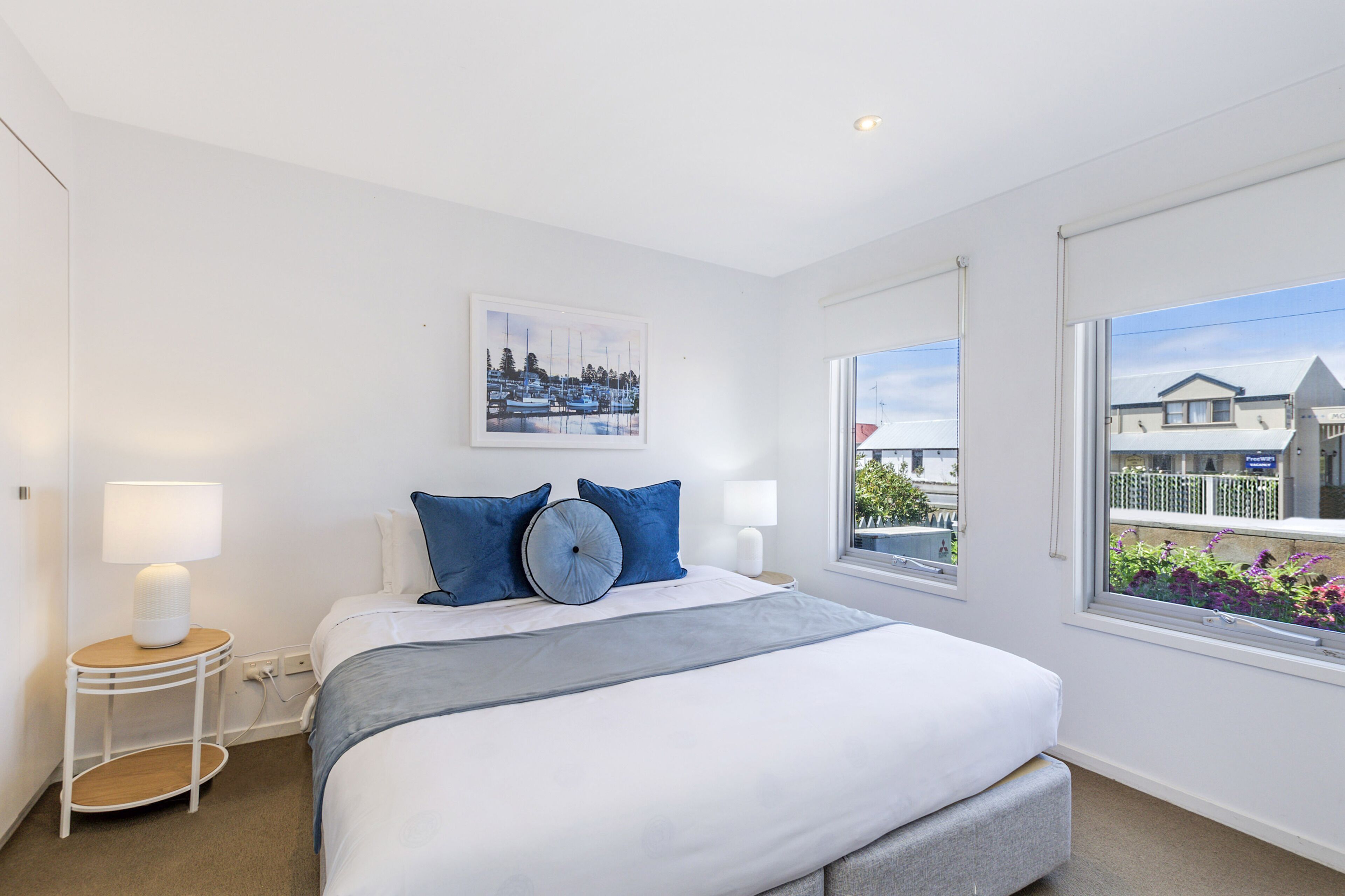 A modern hotel room with a large bed, two bedside tables, a window with a view of buildings, and a painting on the wall.