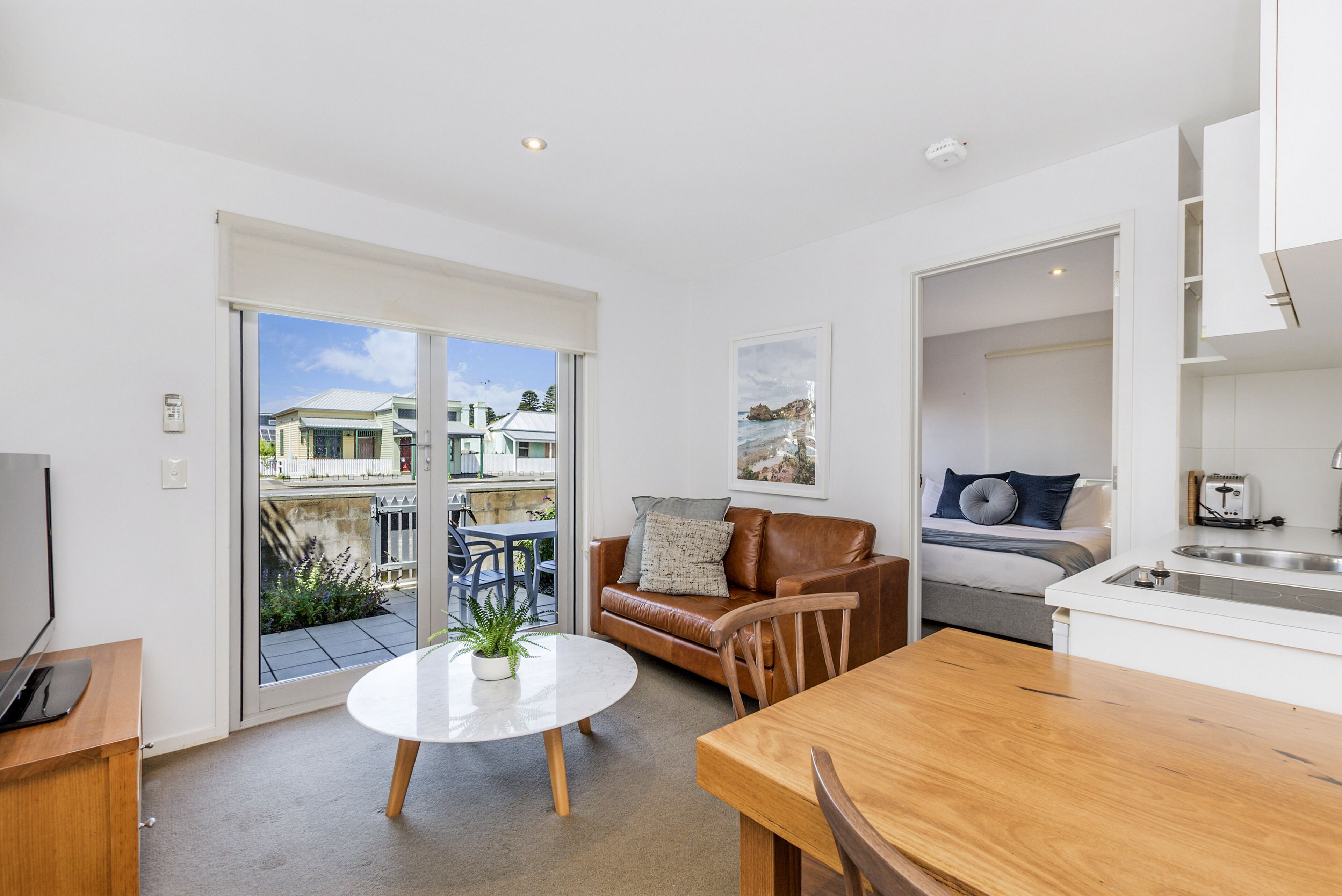 A modern apartment with a dining area, a living room with a brown leather sofa, a kitchen with a white countertop, and a sliding glass door leading to a balcony with a view of other buildings.