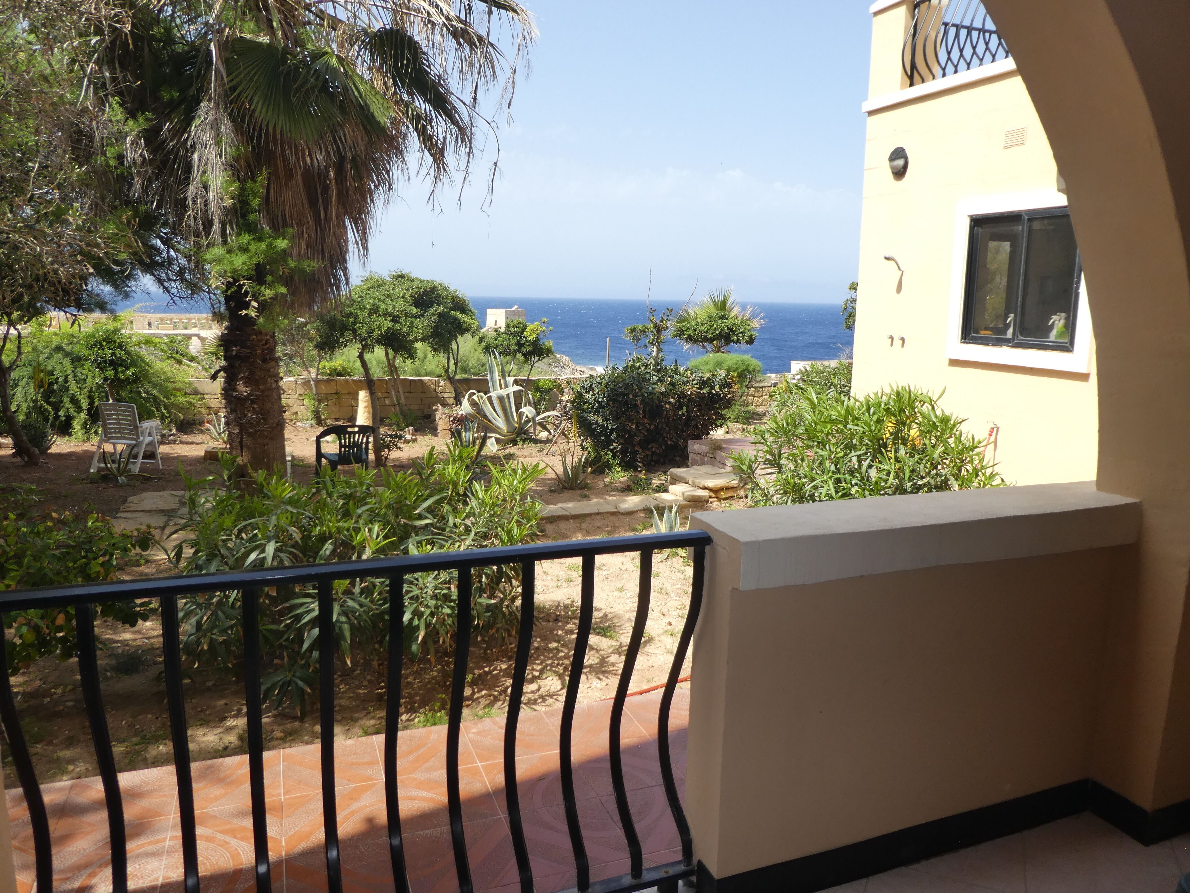 A balcony with a view of the sea, surrounded by greenery and a stone wall.