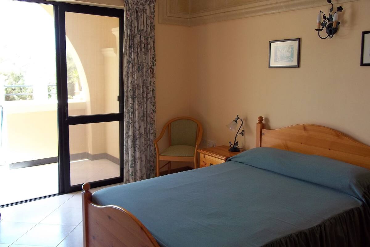 Une chambre à coucher avec un lit, une chaise, une table de chevet, une lampe et une fenêtre avec des rideaux.