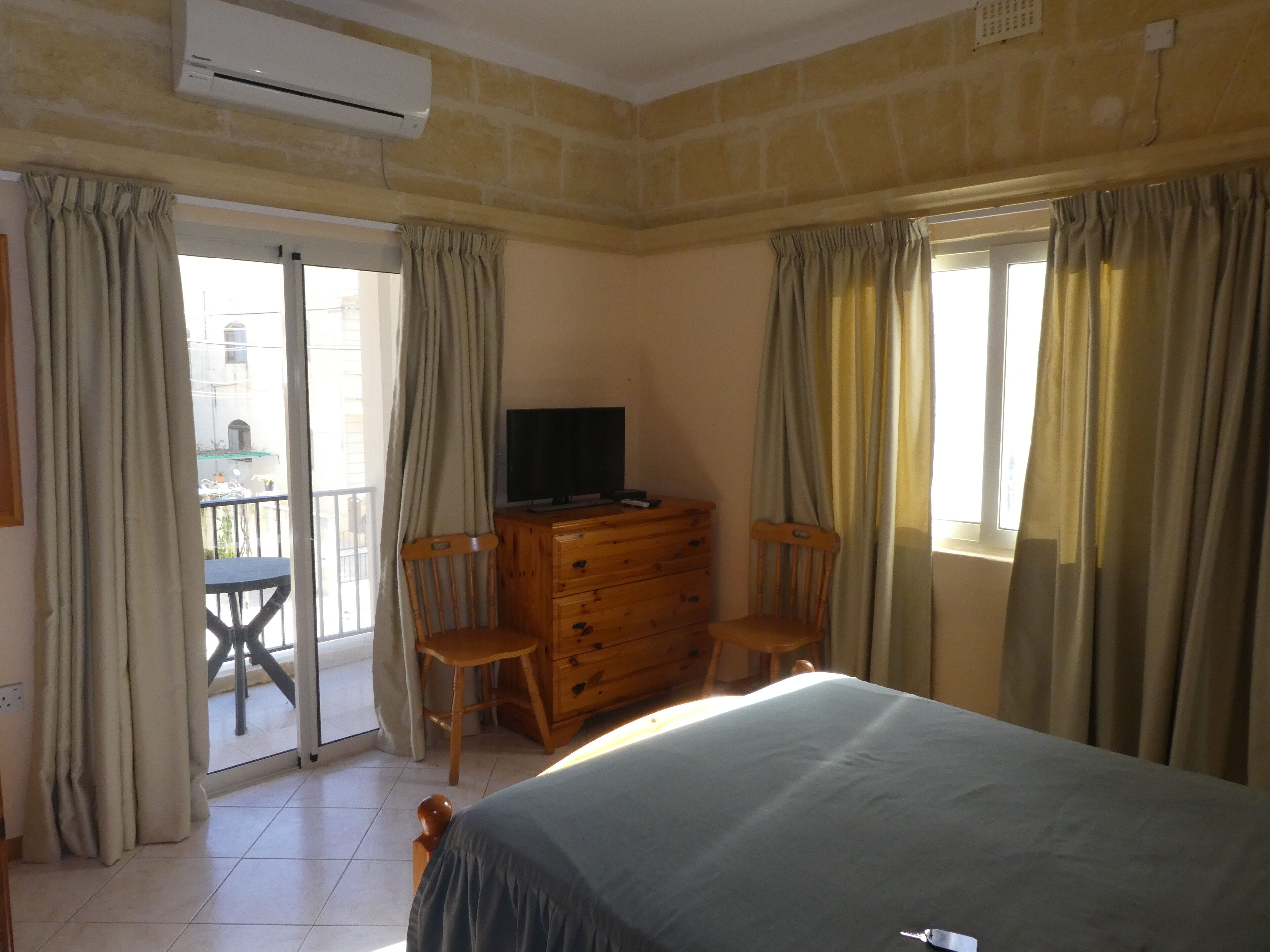 A bedroom with a bed, a wooden dresser, a television on top, and a balcony with a table and chairs.