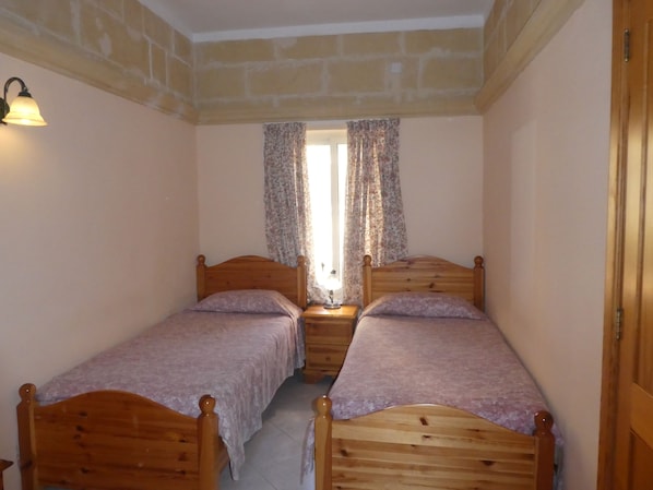 A small, cozy room with two wooden beds, a nightstand with a lamp, and a window with floral curtains.