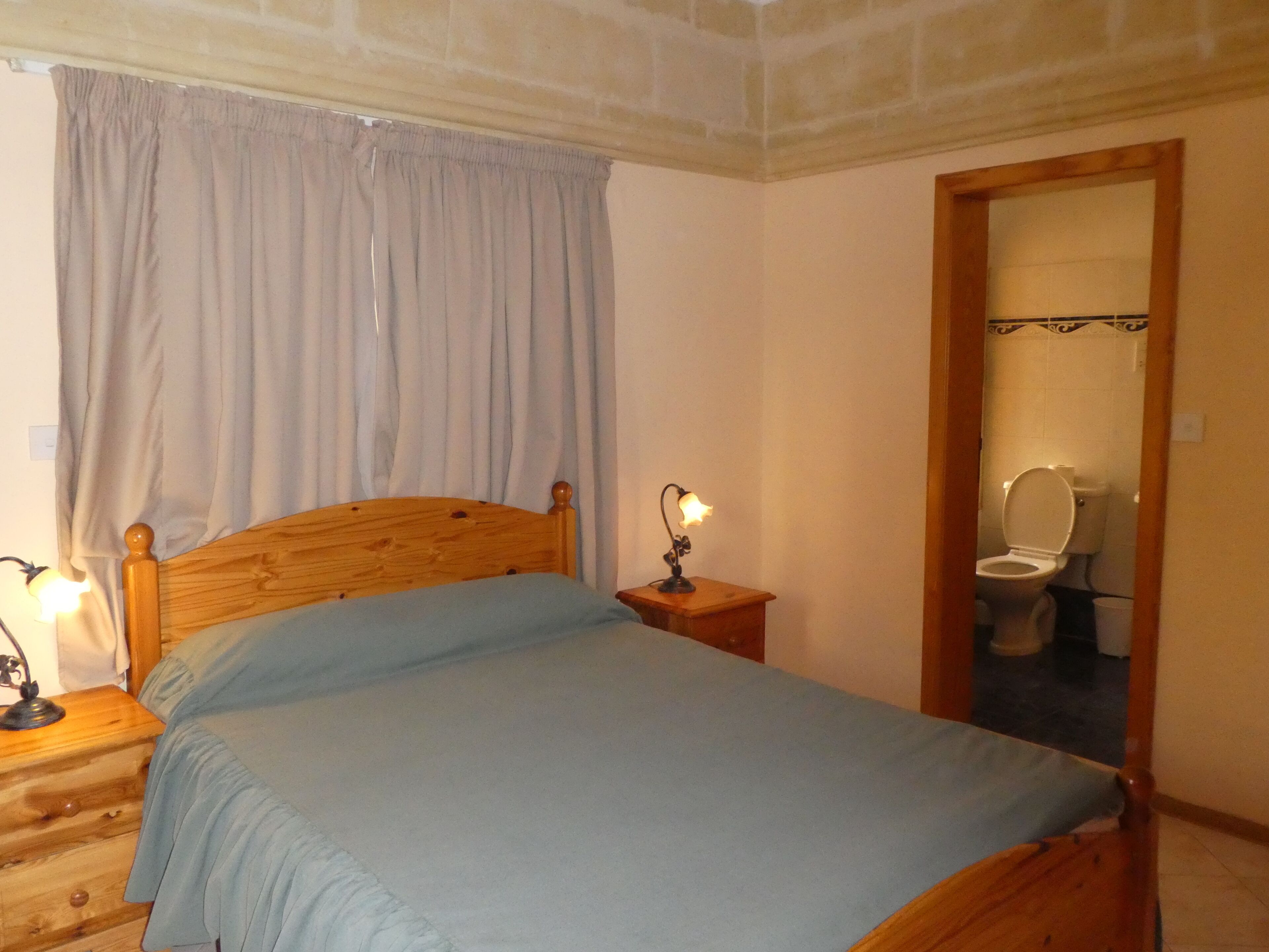 Un dormitorio con cama de madera, una mesita de noche con lámpara, una ventana con cortinas y un baño visible a través de una puerta abierta.