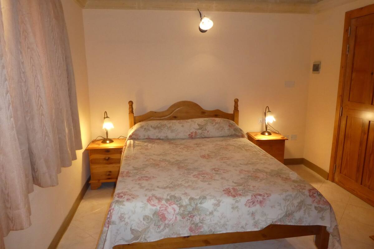 Une chambre avec un lit en bois, deux tables de chevet avec des lampes, une armoire en bois et un rideau sur le côté gauche.