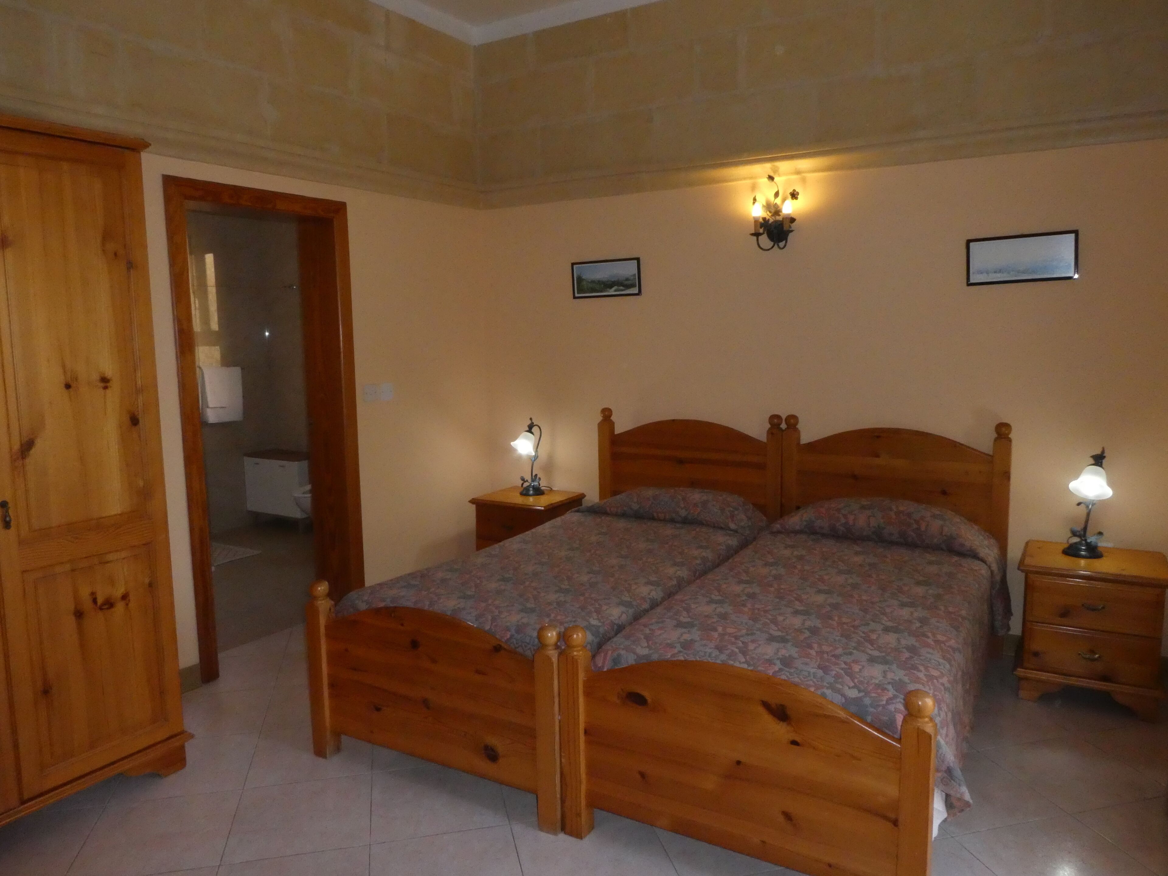 A bedroom with a wooden bed, two bedside tables with lamps, a wooden wardrobe, and a door leading to another room.