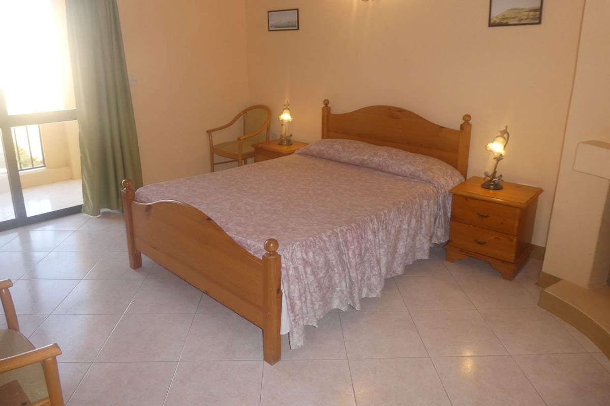 Une chambre à coucher avec un lit en bois, deux tables de chevet avec des lampes, une chaise et une vue sur l’extérieur par une grande fenêtre.
