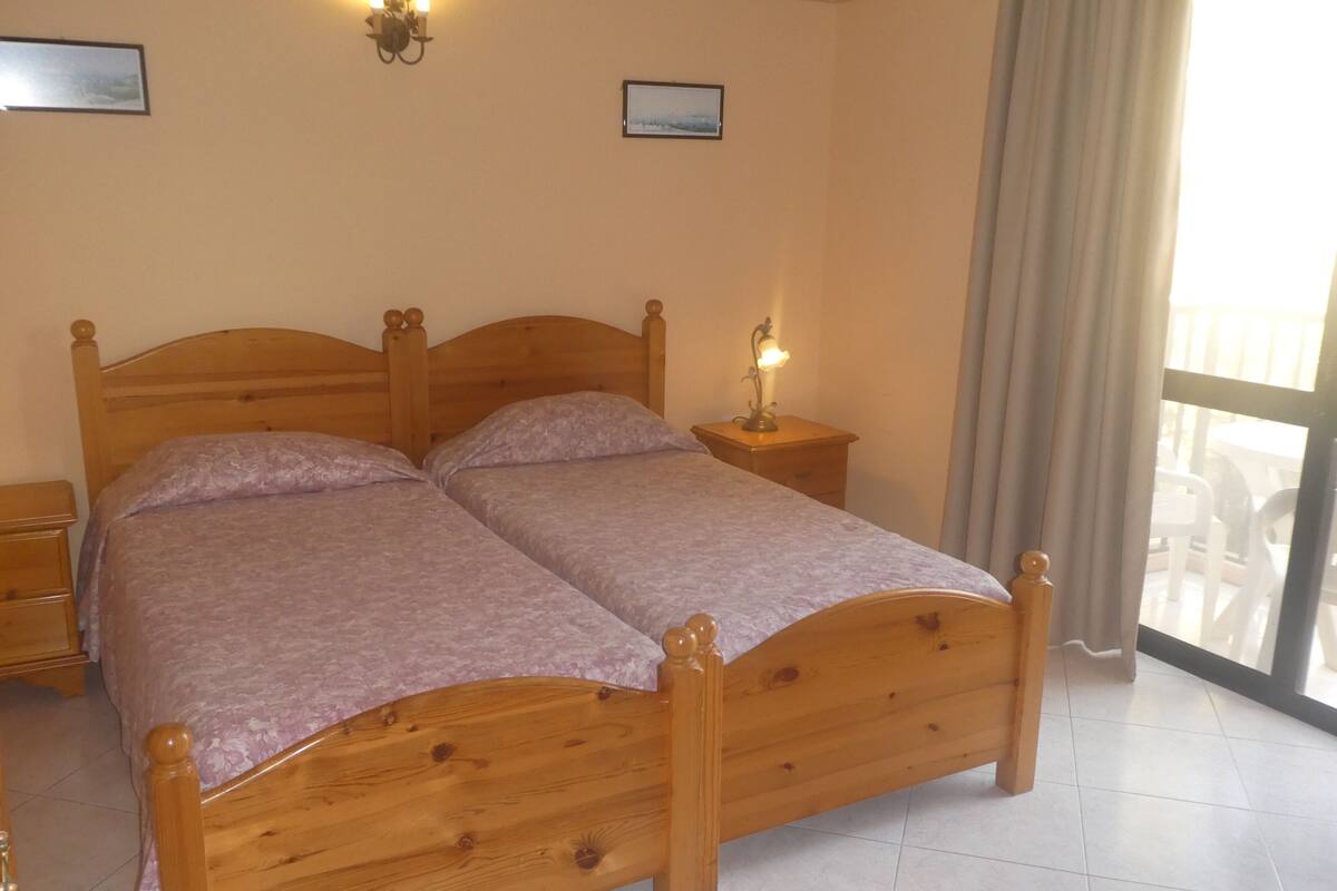Un lit double en bois, avec deux tables de chevet, chacune équipée d’une lampe, dans une pièce dotée d’une grande fenêtre et de rideaux.