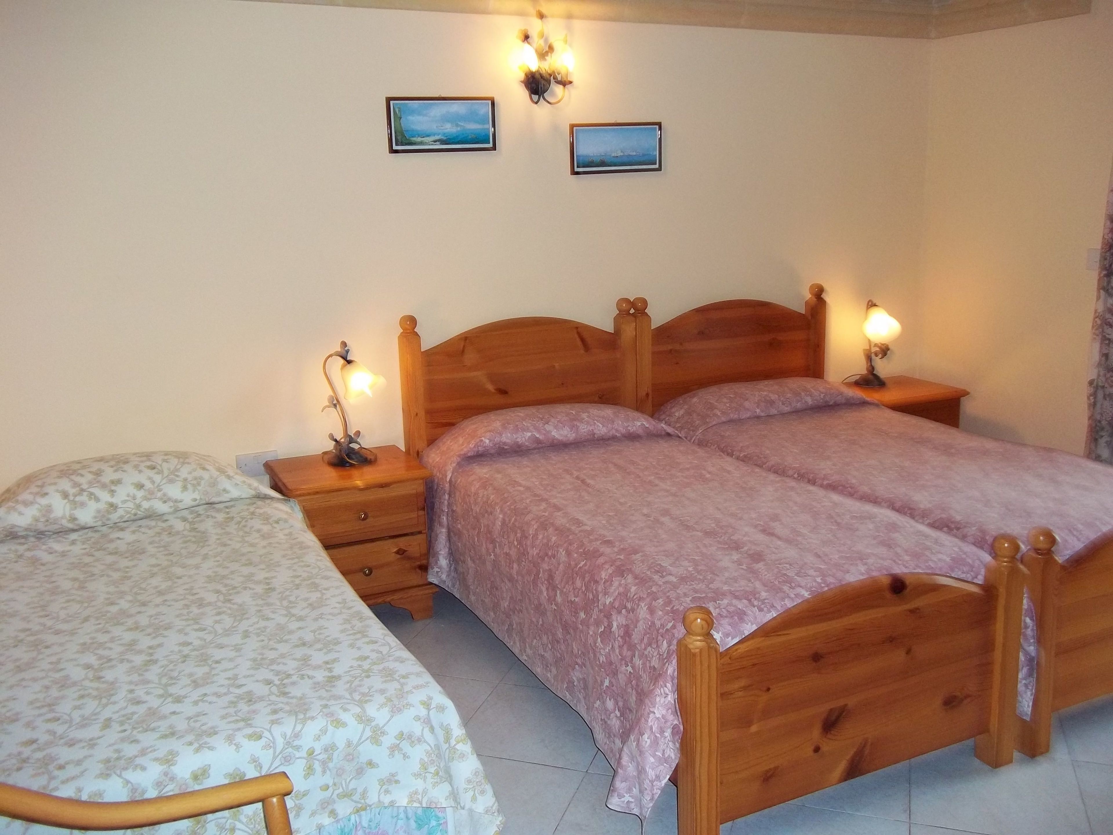 Ein Schlafzimmer mit zwei hölzernen Betten, jeweils mit einem Nachttisch und einer Lampe. Darüber hängen zwei gerahmte Bilder an der Wand.