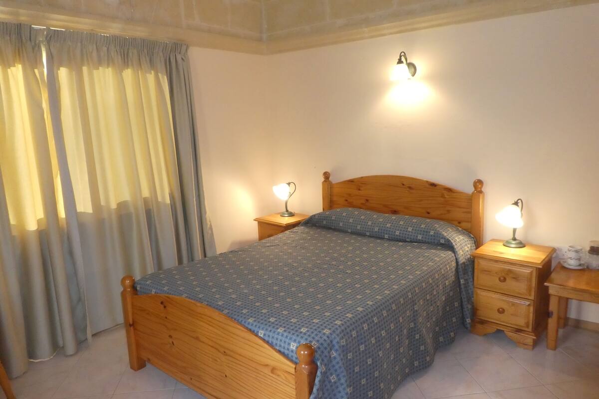 Une chambre à coucher avec un lit en bois, deux tables de chevet avec des lampes, une fenêtre avec des rideaux et un luminaire fixé au mur.