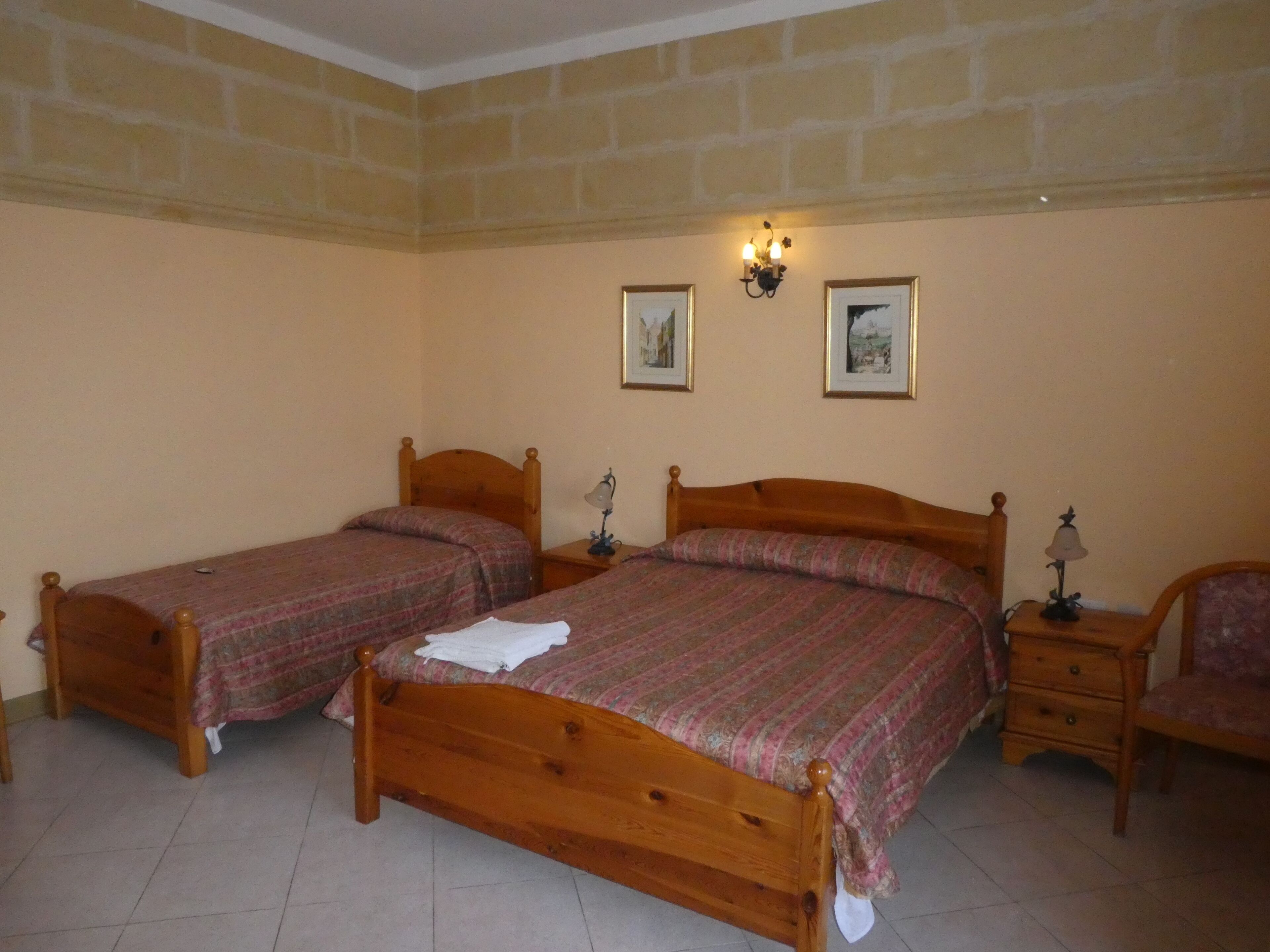 A bedroom with two wooden beds, a nightstand with a lamp, a chair, and two framed pictures on the wall.