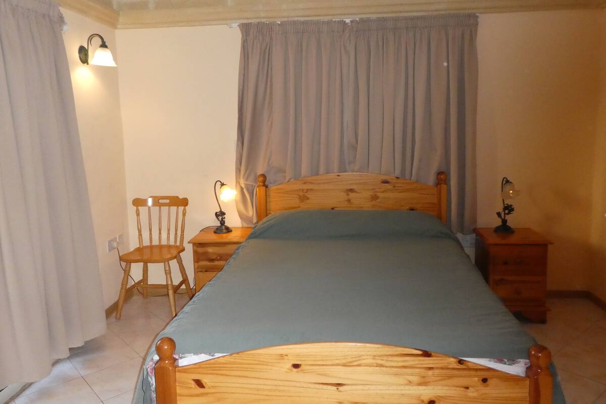 Une chambre à coucher avec un lit en bois, deux tables de chevet avec des lampes, une chaise et une fenêtre avec des rideaux.