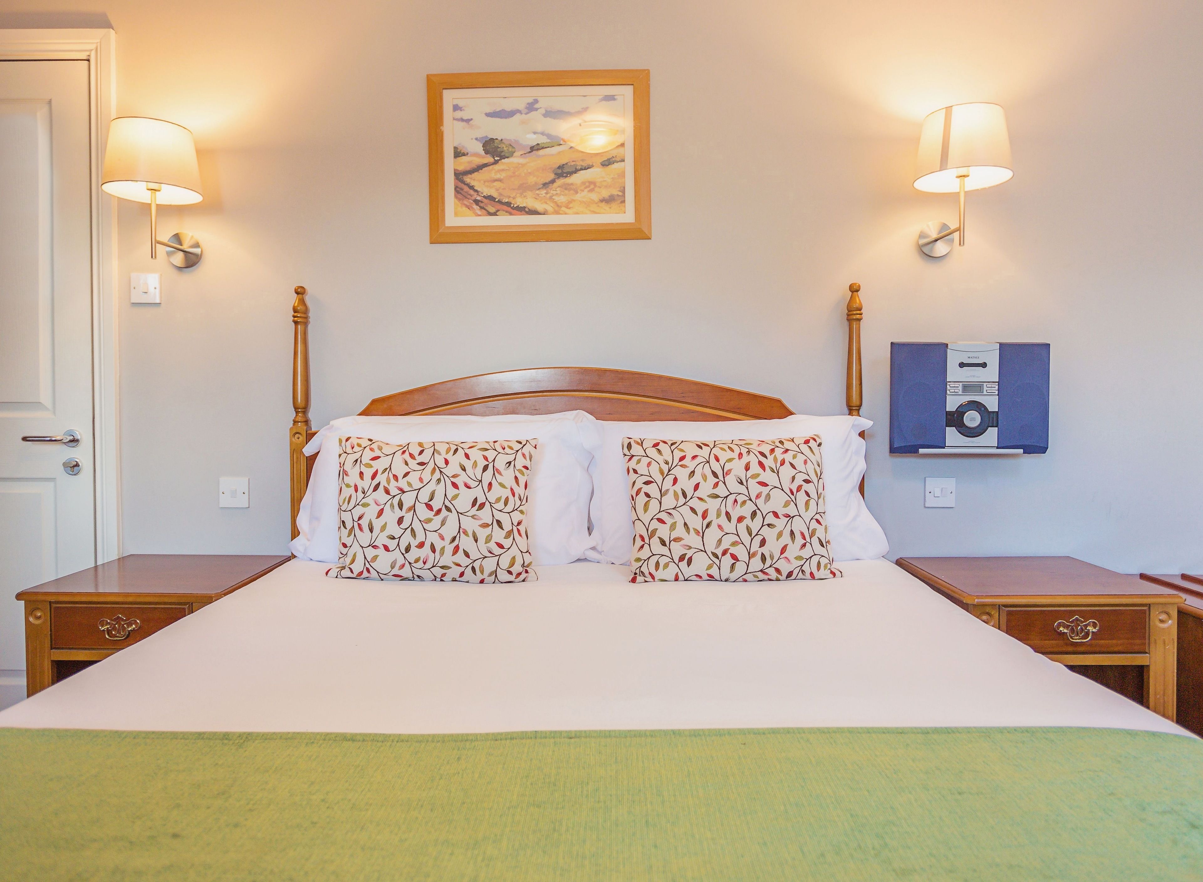 A neatly made bed with two pillows, a bedside table, a wall-mounted lamp, and a framed picture above the bed.