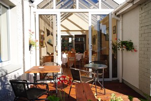 Une terrasse extérieure avec des tables et des chaises en bois, une porte vitrée donnant sur l’intérieur et des plantes en pot.