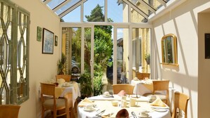 Une véranda avec un toit en verre, des tables à manger préparées pour deux personnes et une vue sur le jardin.