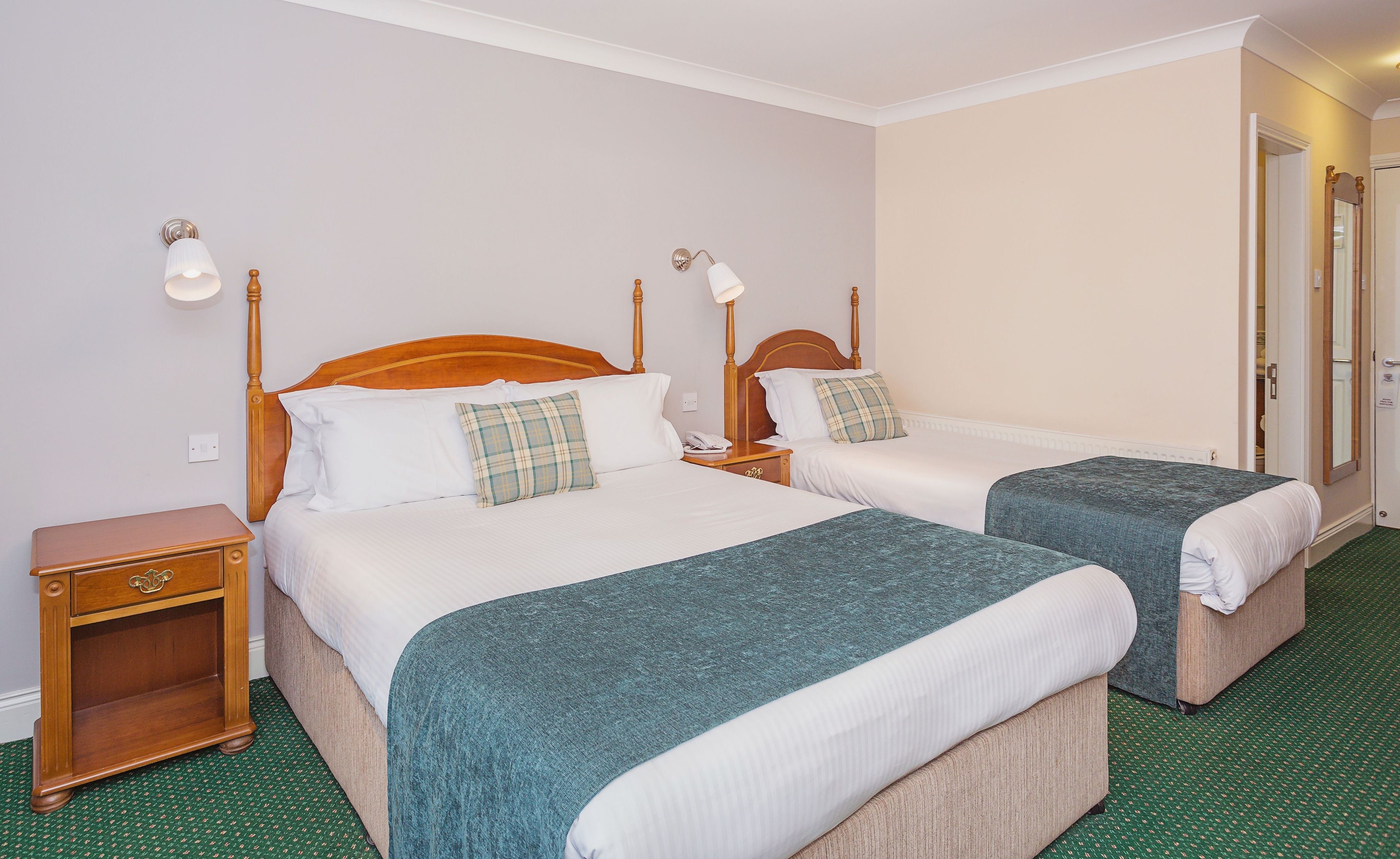 A hotel room with two single beds, each with a wooden headboard, white bedding, and a green throw blanket. There are bedside tables with lamps, a wooden nightstand, and a green carpeted floor.
