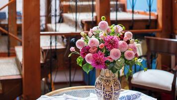 Una habitación con una escalera de madera, una mesa redonda con mantel azul y blanco estampado y un jarrón con flores rosadas.