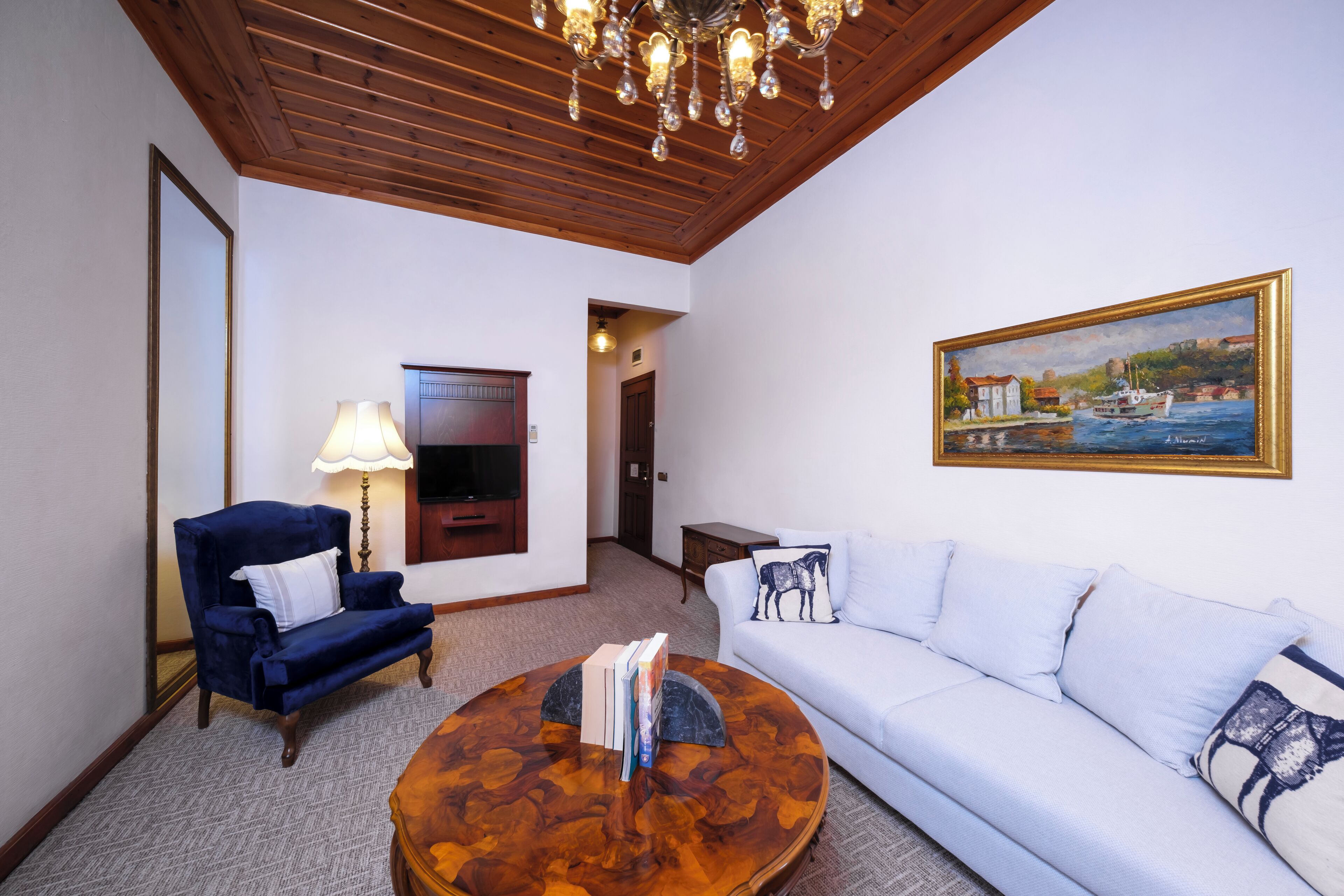 A living room with a blue armchair, white sofa, and a round coffee table with books. There is a painting on the wall and a chandelier.