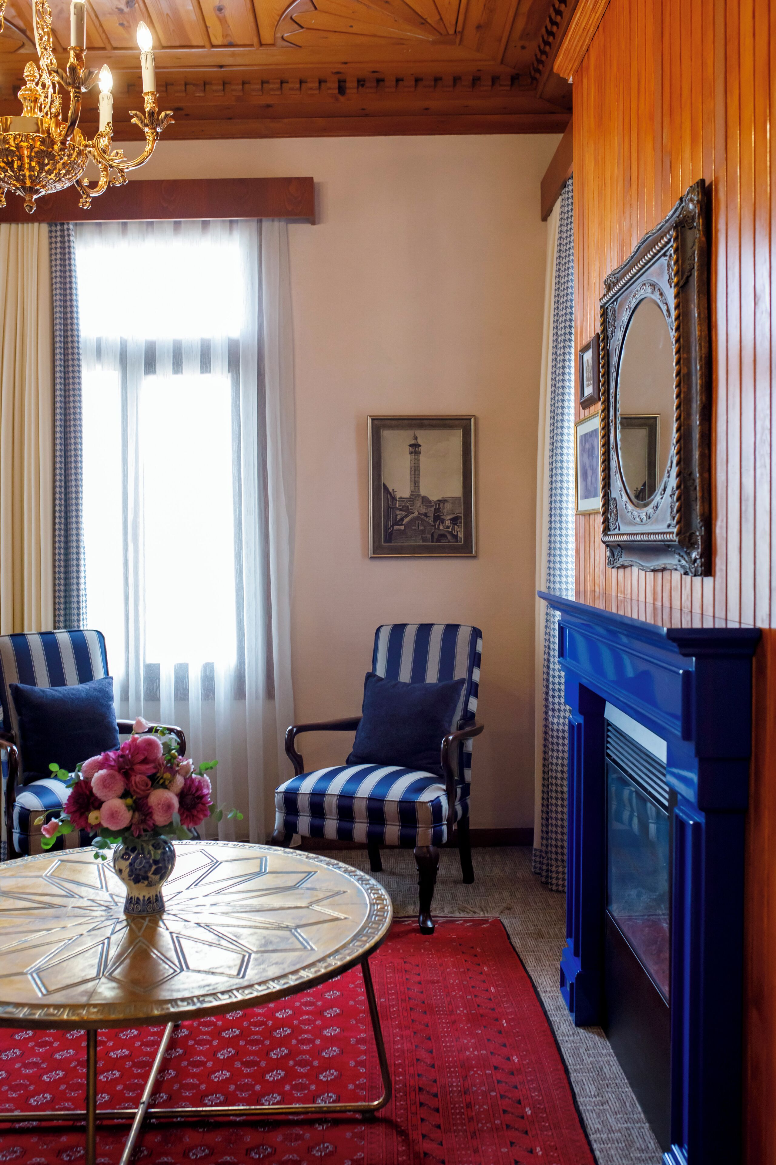 Una habitación con una mesa redonda, dos sillas a rayas, un florero con flores, una chimenea y un cuadro en la pared.