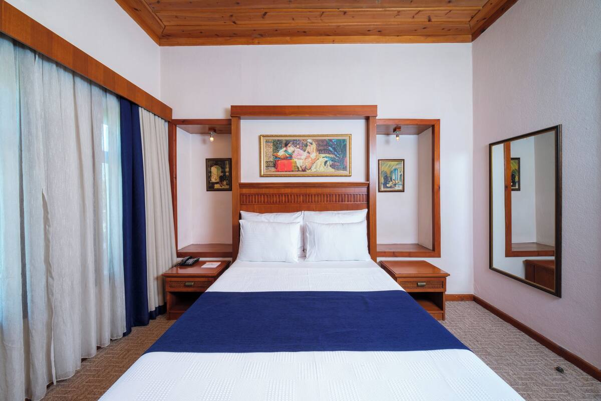 Une chambre d’hôtel avec un grand lit, une tête de lit en bois, deux tables de chevet, une commode, un tableau au-dessus du lit, un miroir au mur et une fenêtre avec des rideaux.