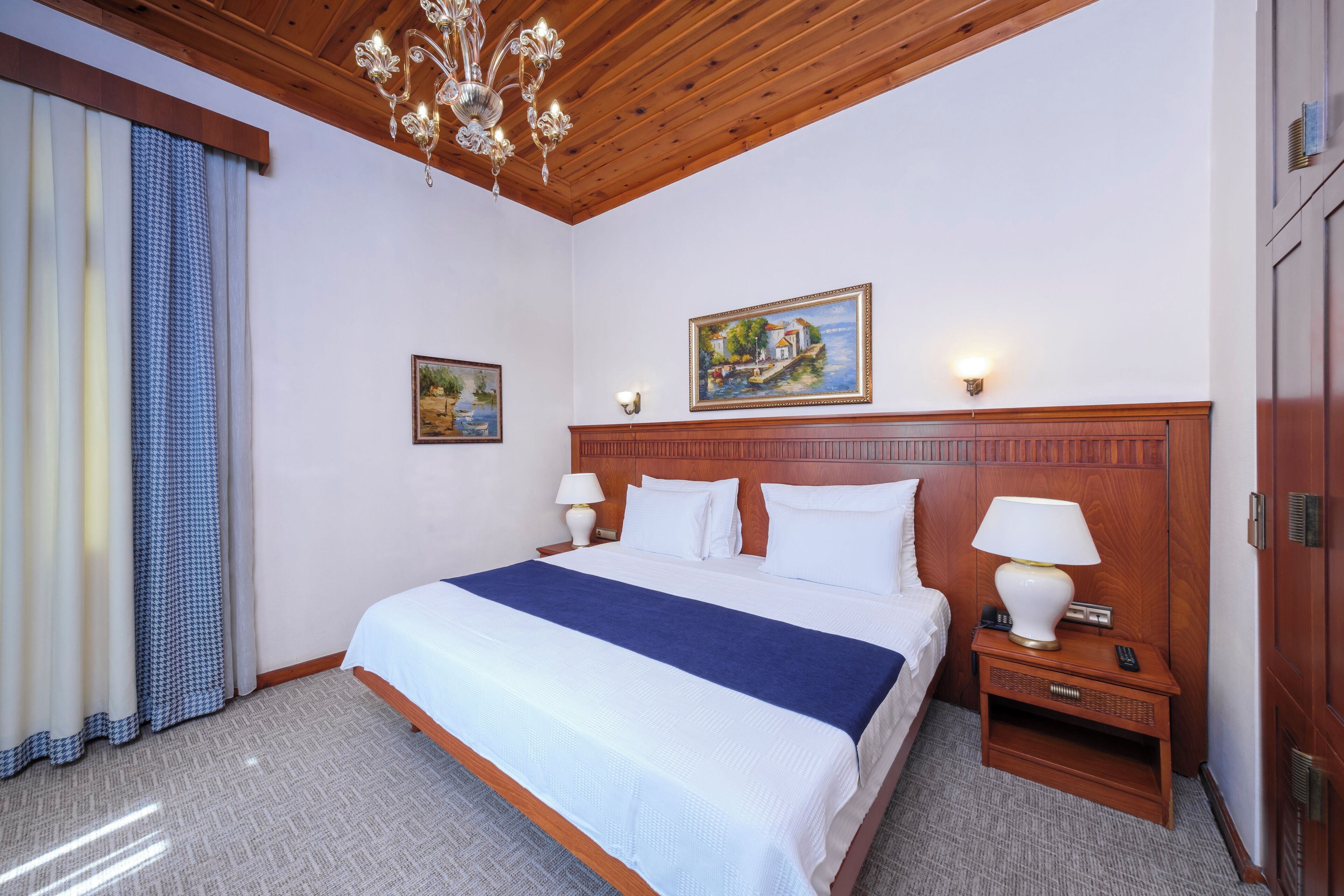 Een hotelkamer met een houten plafond, een groot bed met een blauwe deken, twee nachtkastjes met lampen, en een schilderij aan de muur.
