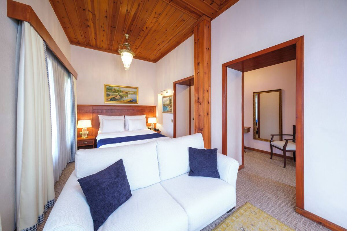 Une chambre d’hôtel avec un plafond en bois, un canapé blanc, un lit recouvert d’une couverture bleue et un tableau accroché au mur.