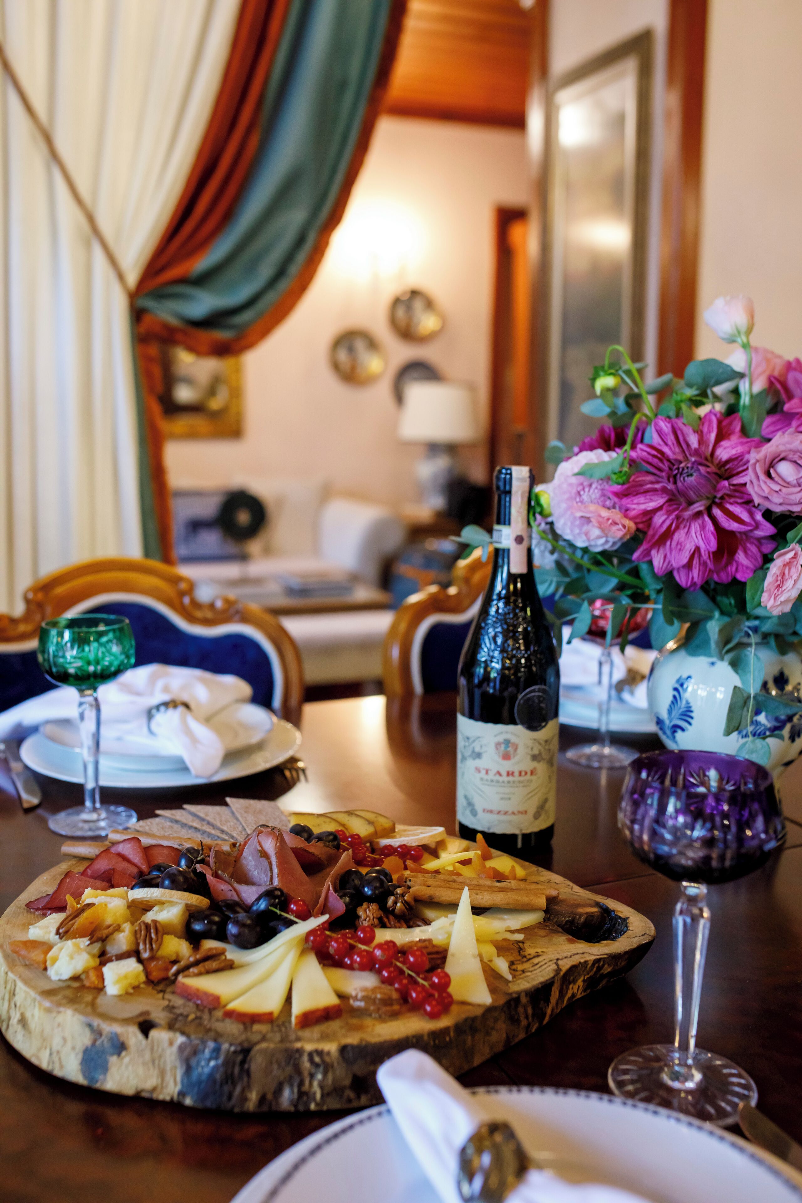 Una mesa con una tabla de quesos, una botella de vino y un florero decorativo.