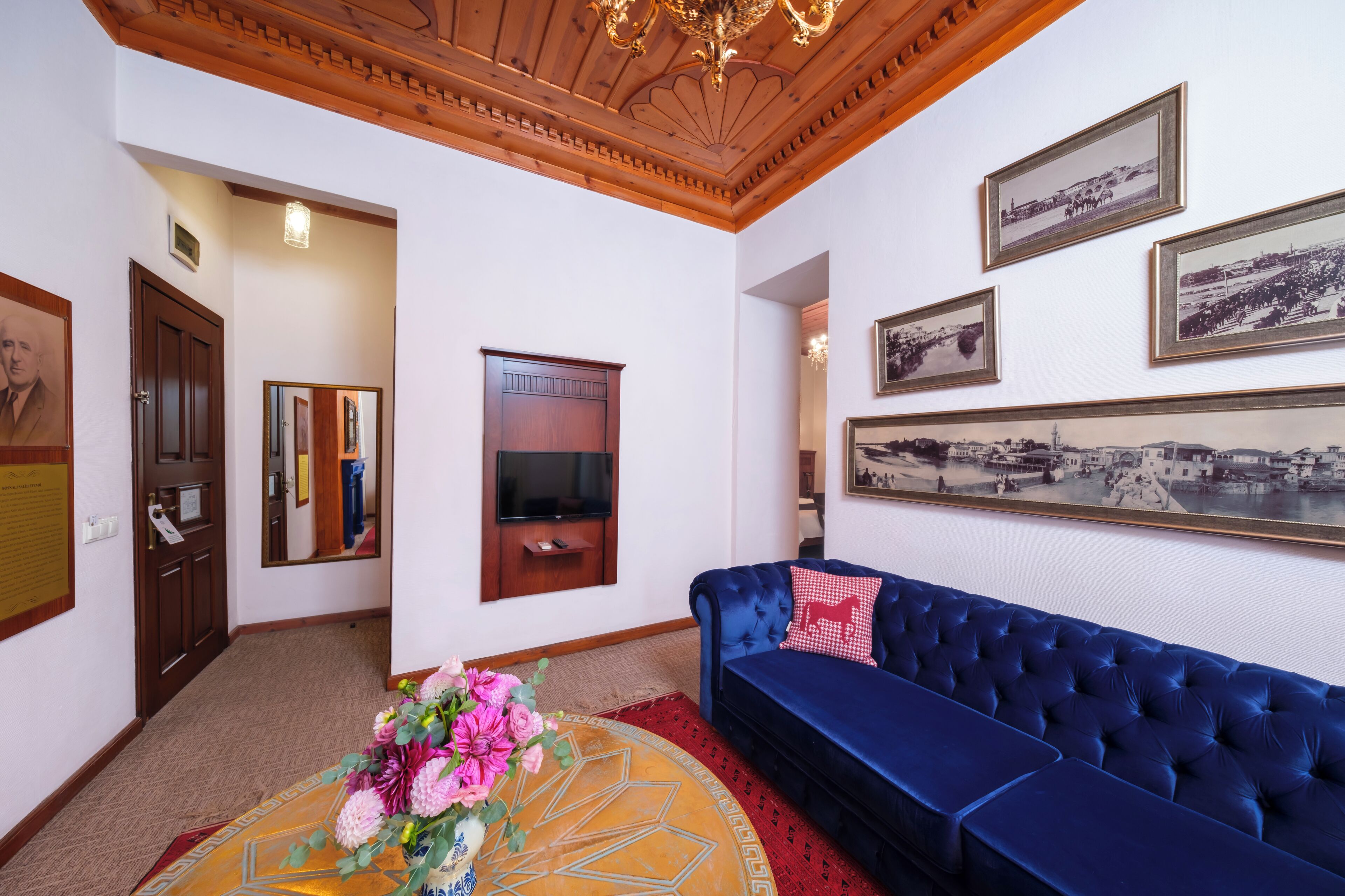 A room with a blue sofa, a wooden ceiling, framed pictures on the wall, and a television mounted on the wall.