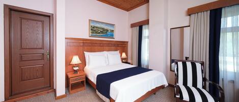 A hotel room with a wooden door, a bed with white bedding, a nightstand with a lamp, a striped armchair, and a painting on the wall.