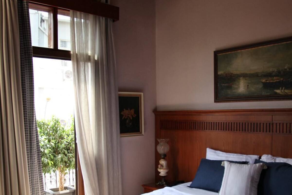 Une chambre d’hôtel avec un grand lit, une tête de lit en bois, un tableau au mur, une fenêtre avec des rideaux et une plante en pot.