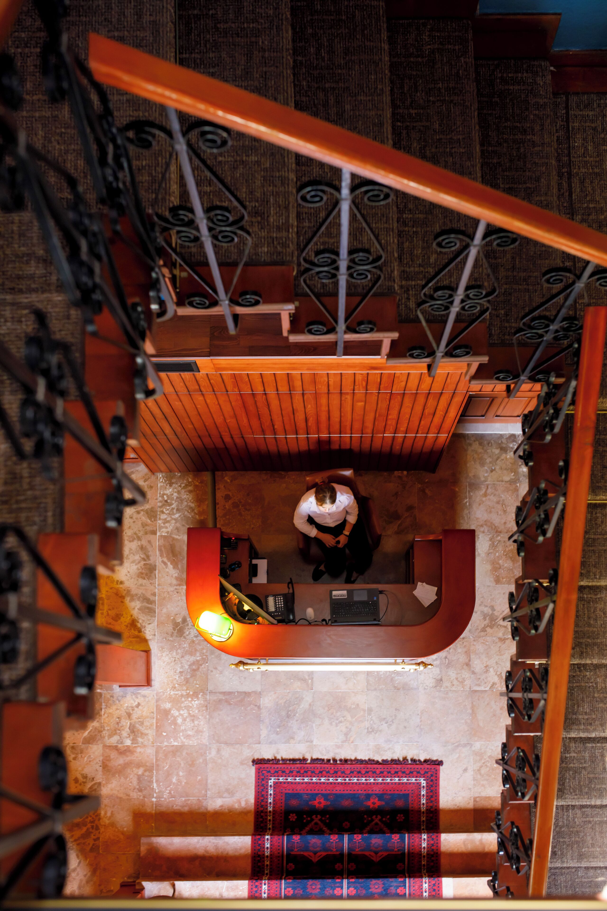 A staircase with a wooden handrail and metal balusters, leading to a landing area with a patterned rug and a desk.