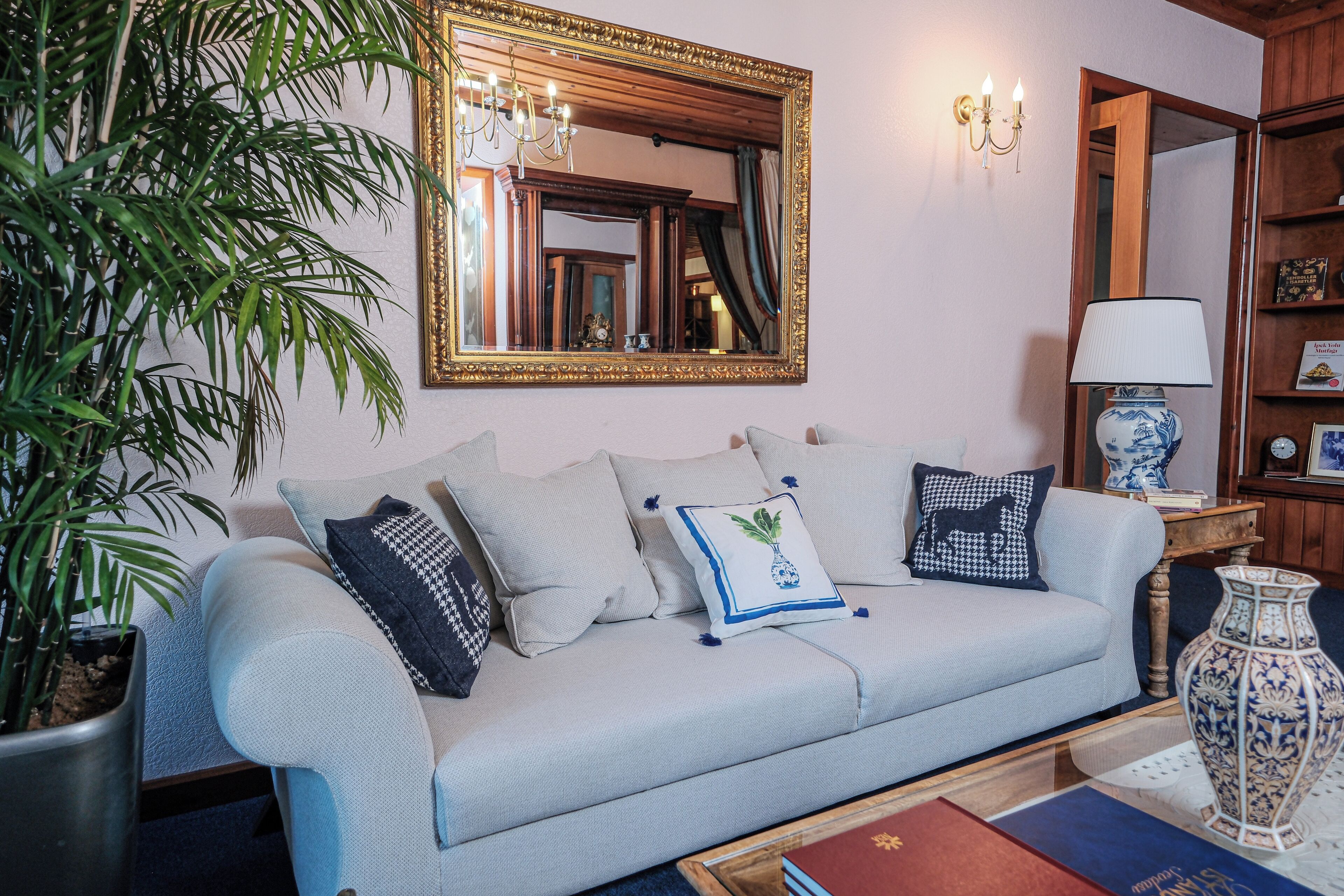 A living room with a light blue sofa, various patterned cushions, a large mirror with a gold frame, a potted plant, and a vase on a side table.