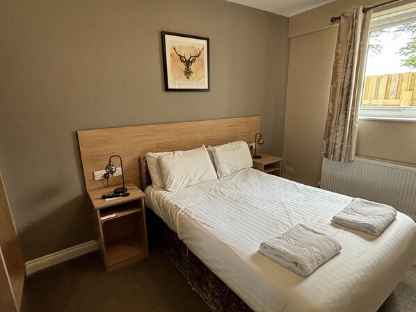 Una habitación de hotel con una cama grande, dos mesitas de noche, una obra de arte con un ciervo enmarcado y un ventanal con cortinas.