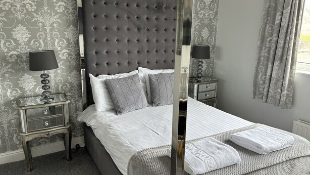 A bedroom with a tufted headboard, a bed with white linens, two bedside tables with lamps, and a window with curtains. - Solway Lodge Hotel (Gretna)