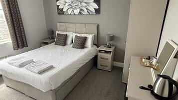 A neatly arranged bedroom with a large bed, two bedside tables, a nightstand, a wall-mounted lamp, a mirror, and a kettle on a cabinet.