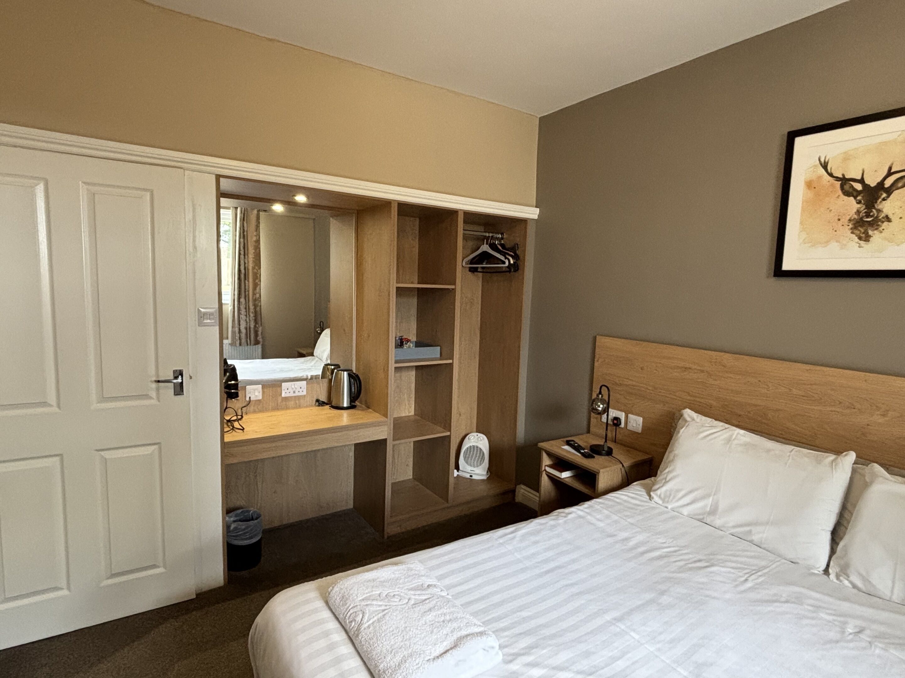 Et hotelværelse med en stor seng, et træhovedgærde, et natbord med en lampe, et lille natbord med en fjernbetjening, et spejl, en træskab og et billede i ramme på væggen.