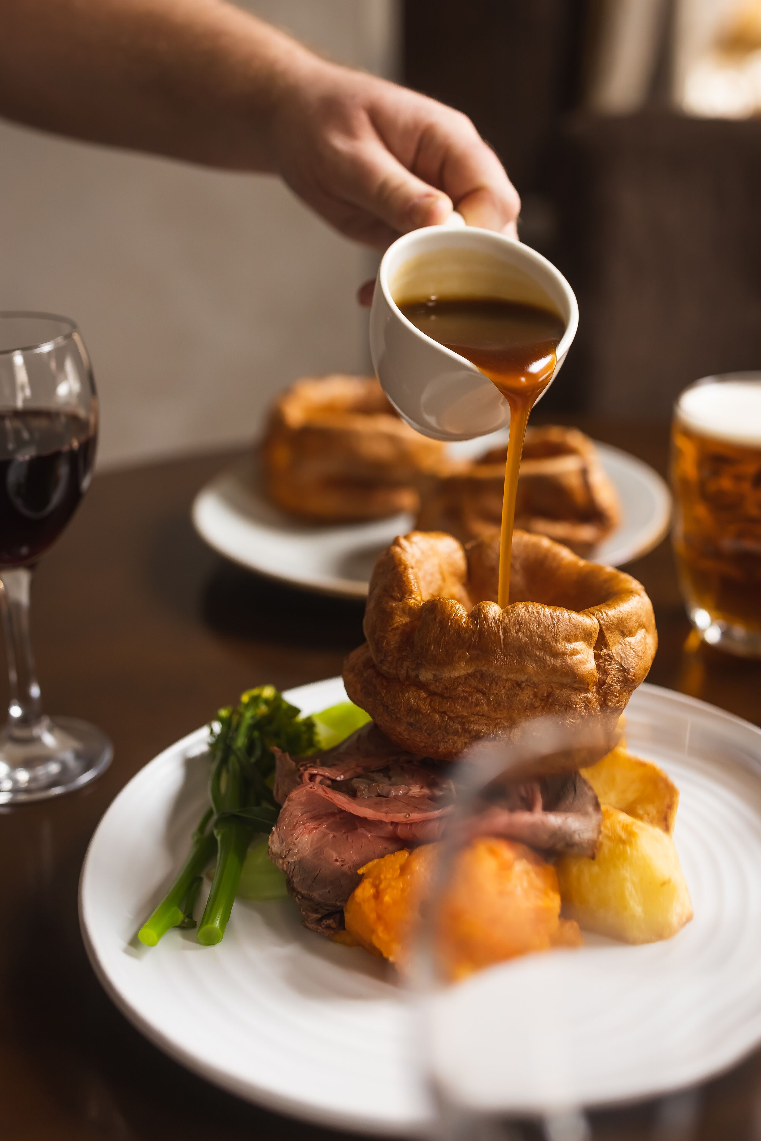 A plate of food with Yorkshire pudding, roast beef, and vegetables, and a hand pouring sauce over it.