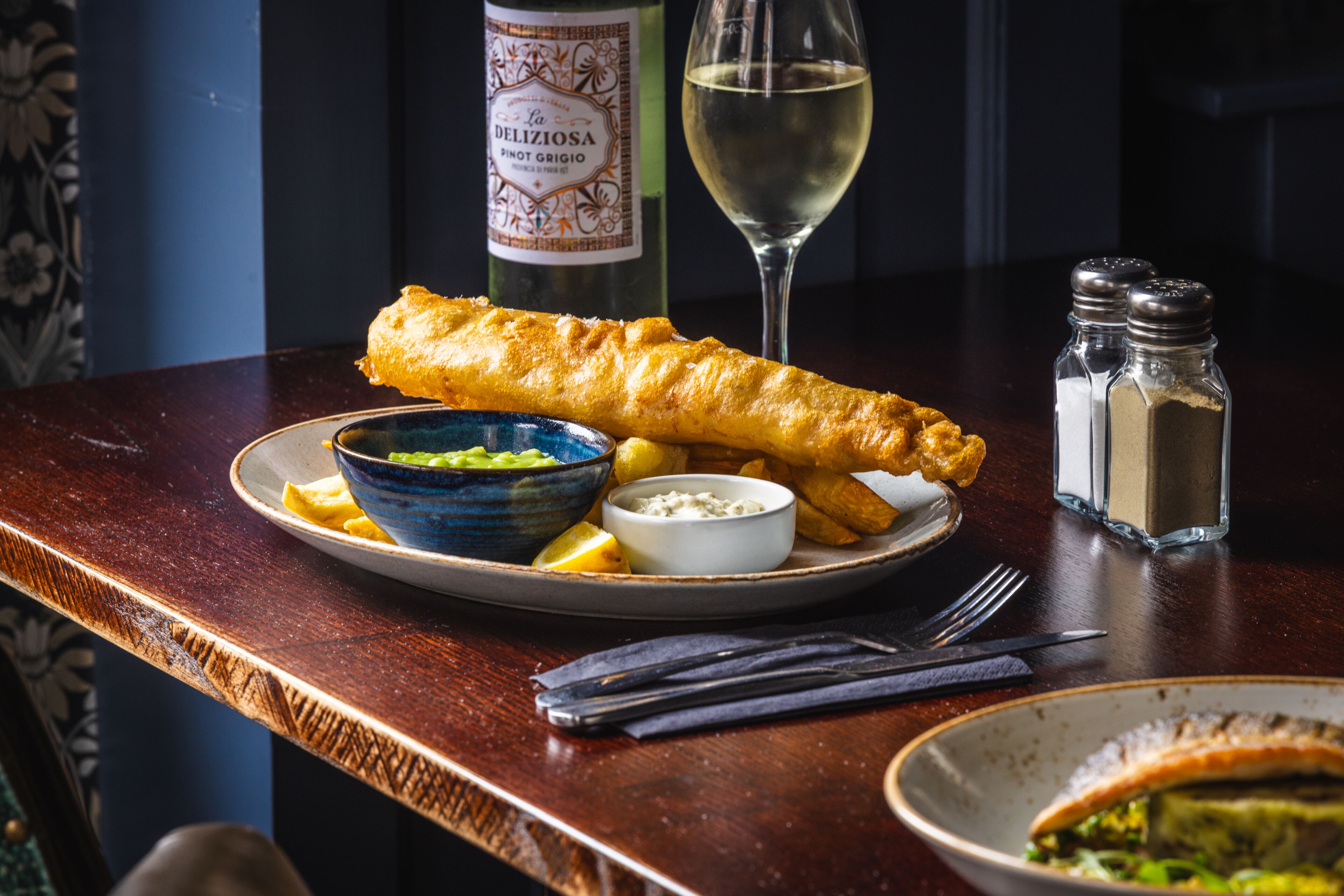 En tallerken med fish and chips, et glas hvidvin og tilbehør.