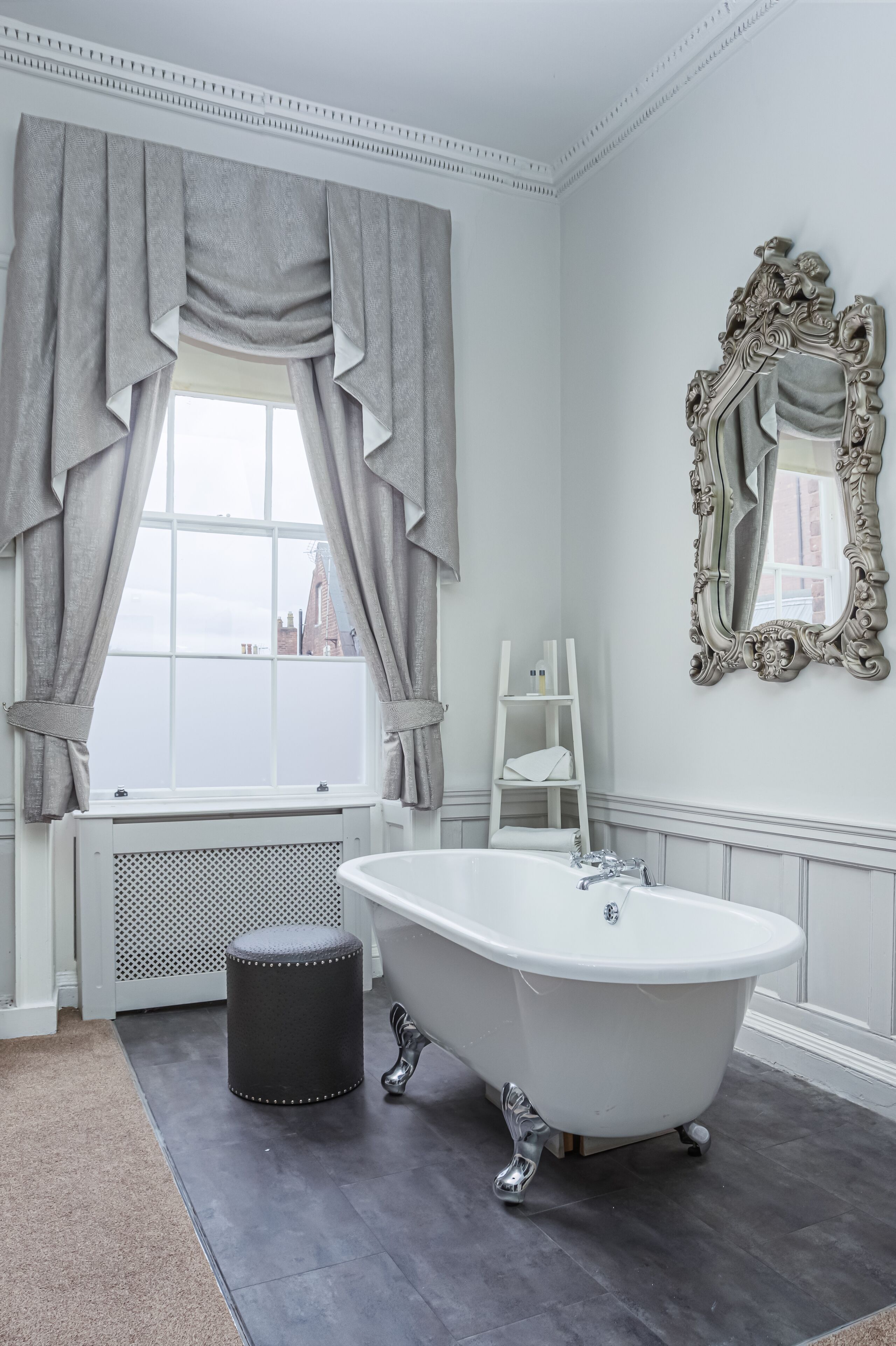 Un bagno con una vasca freestanding, una finestra con tende, un termosifone e uno specchio con cornice decorata.