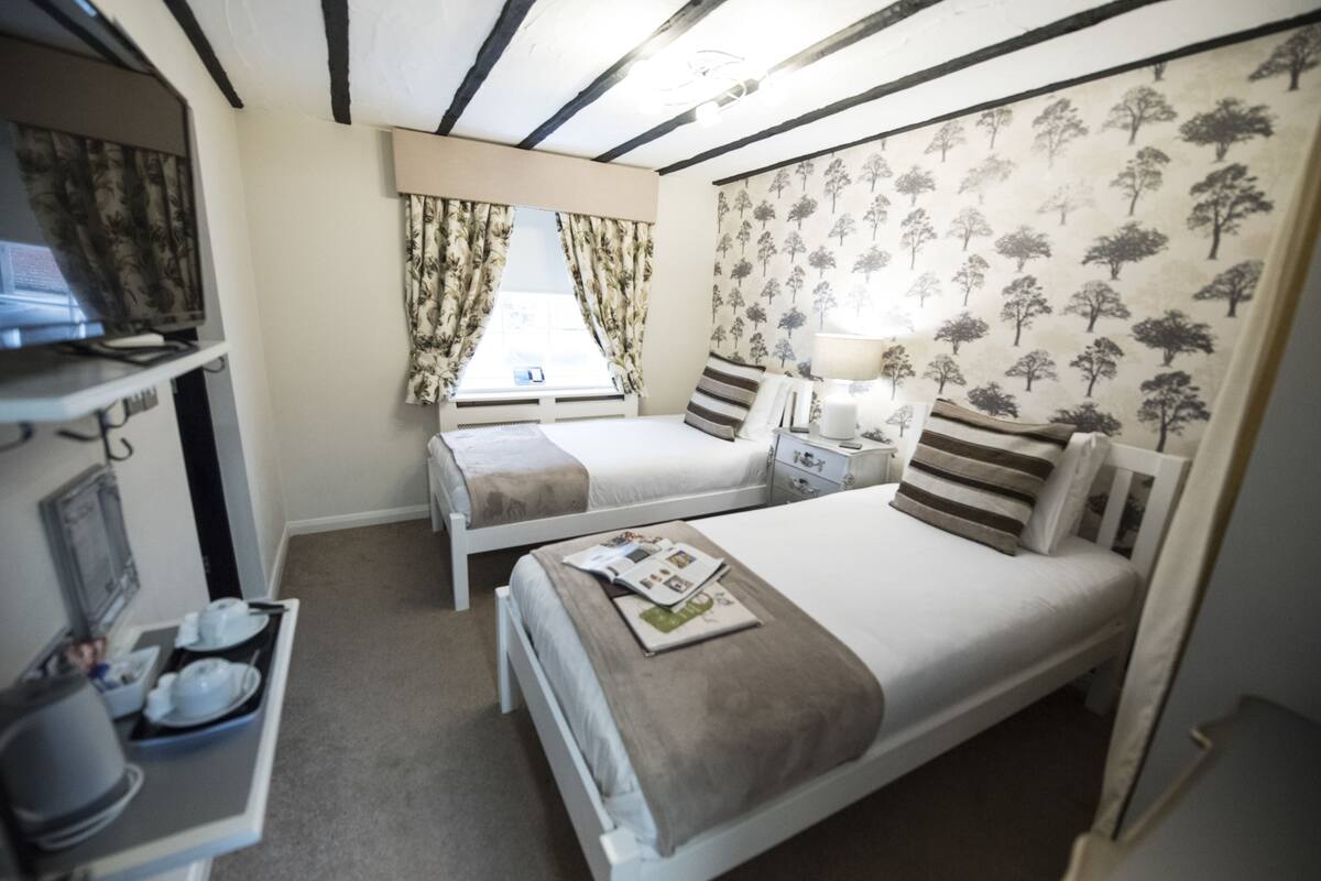 Habitación de hotel con dos camas, un escritorio con computadora, una ventana con cortinas y una pared con estampado de árbol.