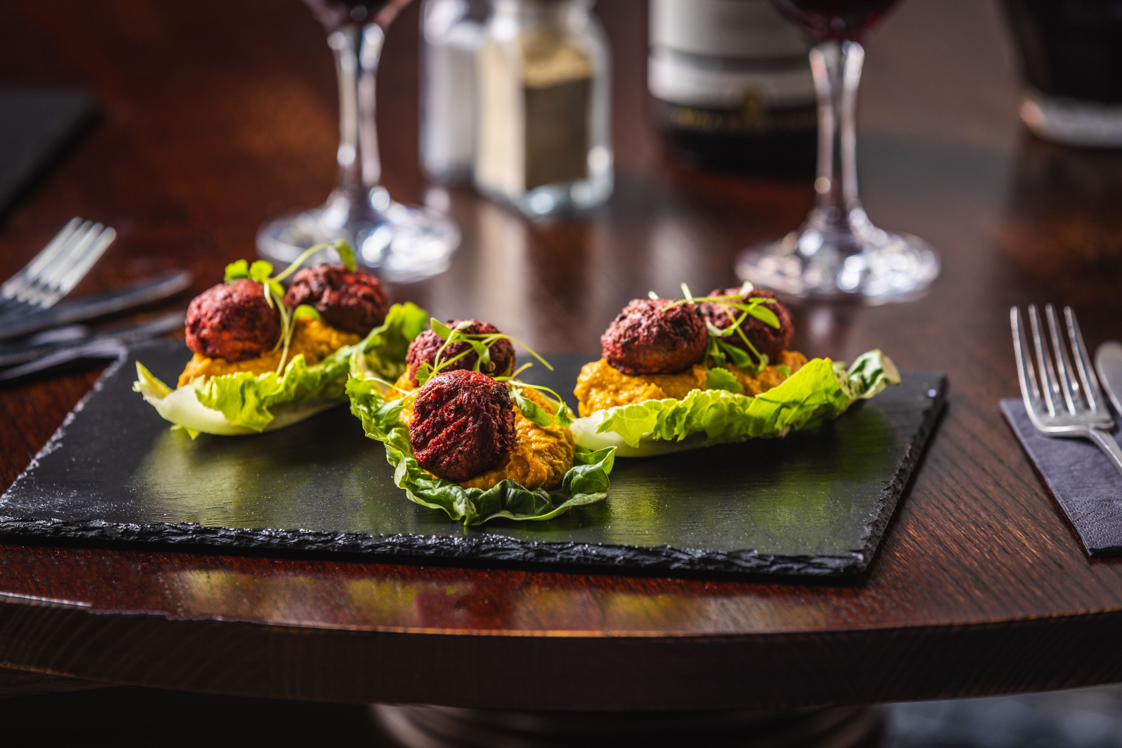 Une table à manger sur laquelle repose un plateau de service en ardoise noire, présentant trois boulettes de viande sur des feuilles de laitue, accompagné de verres à vin et de couverts.