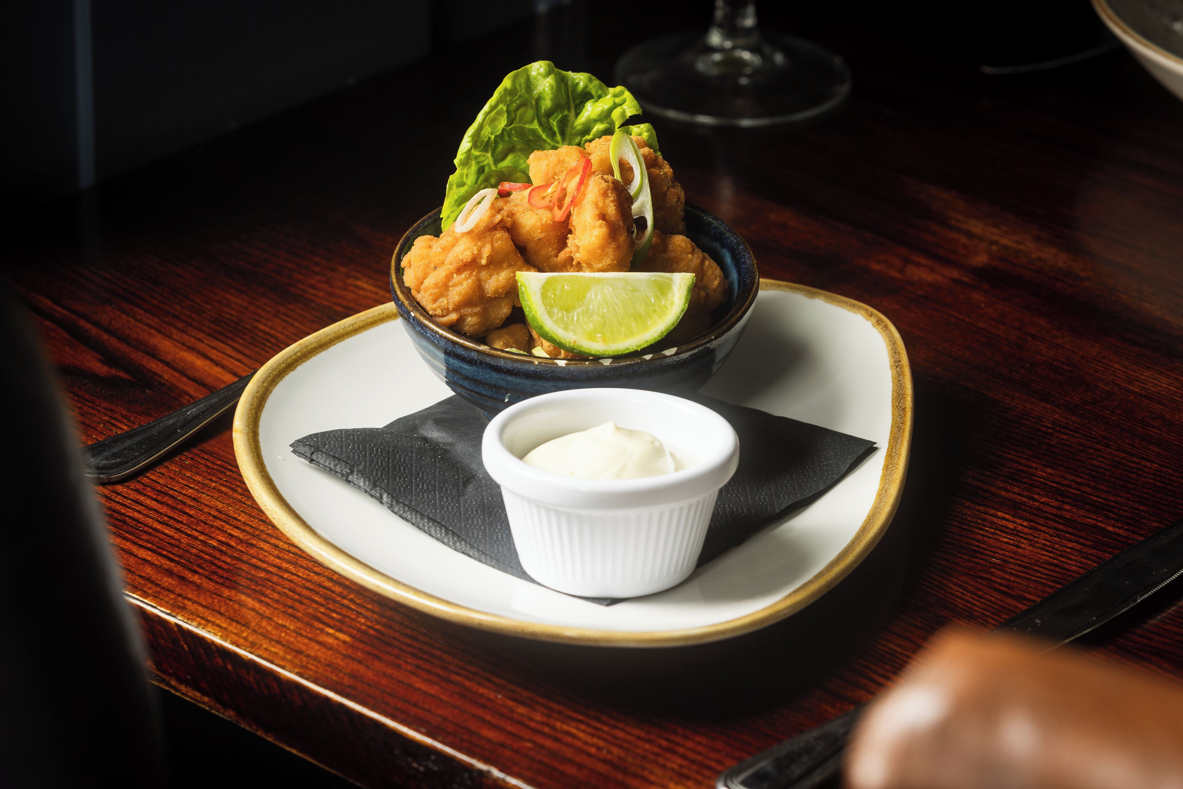 Un plato con comida frita, una cuña de lima y un recipiente pequeño con salsa.
