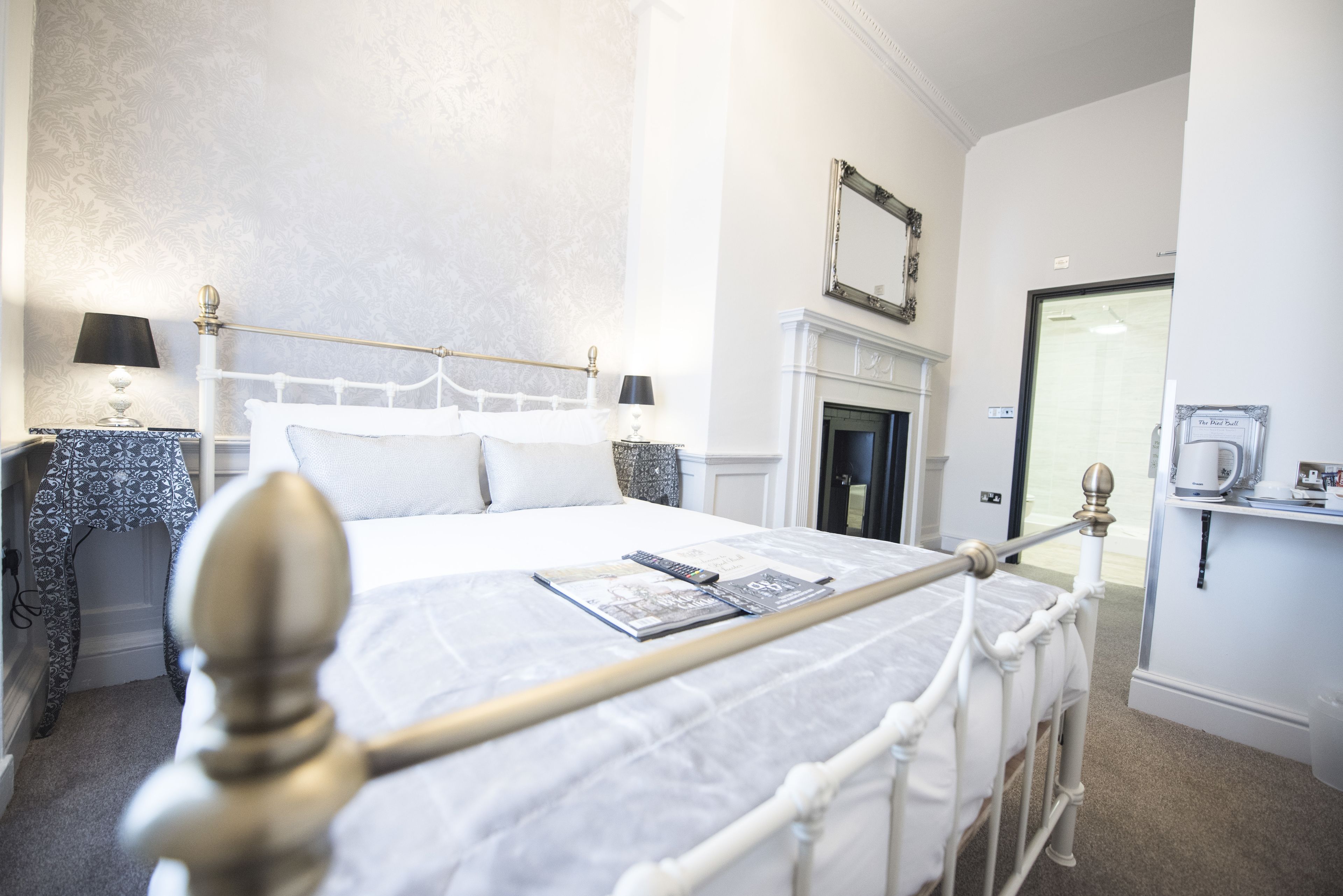 A neatly made bed with a white headboard, a bedside table with a lamp, and a fireplace in the background.