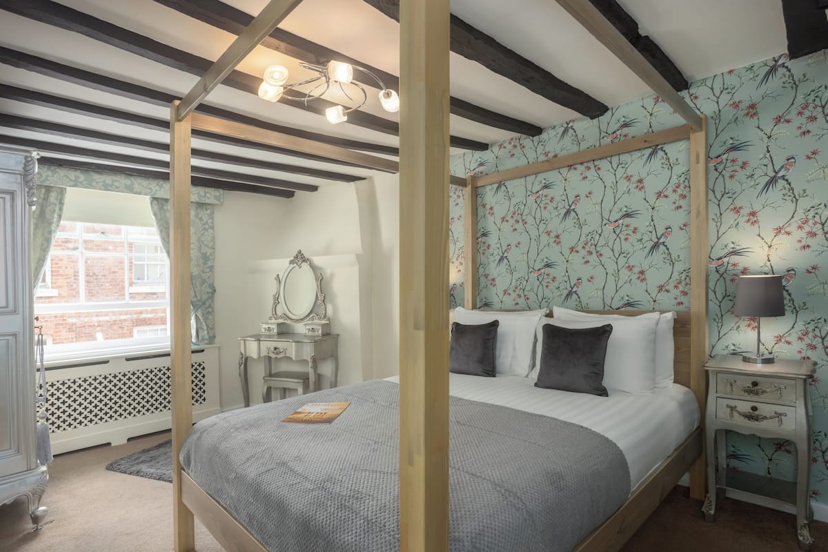 Un dormitorio con cama con dosel, una mesita de noche con lámpara y una ventana con cortinas estampadas.