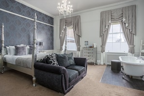A bedroom with a four-poster bed, a dark-colored sofa, a white clawfoot bathtub, and a large window with curtains. - The Pied Bull (Chester)