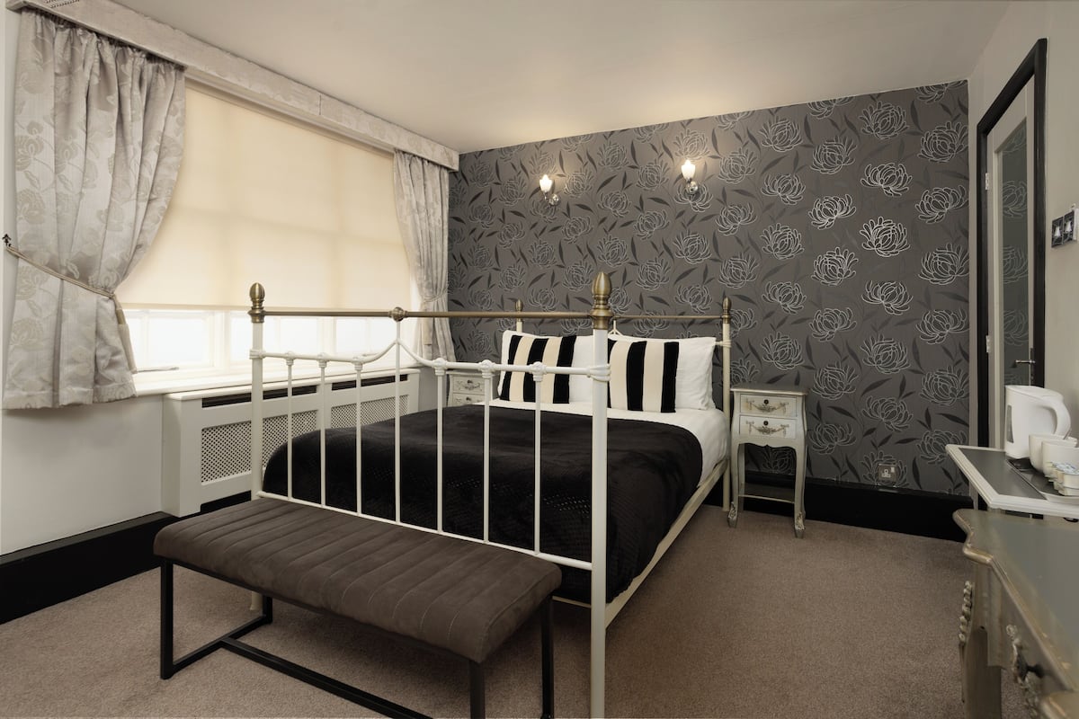 Un dormitorio con estructura de cama de metal blanco, una mesita de noche, un banco, un radiador, una ventana con cortinas y un papel tapiz floral.
