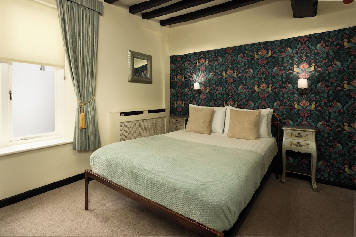 Un dormitorio con cama, mesita de noche, cómoda, ventana con cortinas y pared con estampado floral.