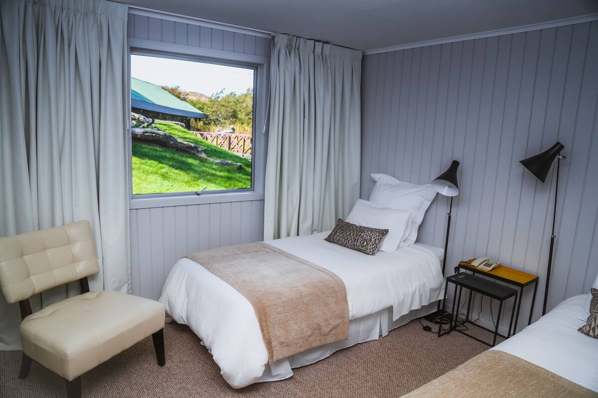Ein ordentlich eingerichtetes Schlafzimmer mit Bett, Sessel, Nachttisch mit Lampe und einem Fenster mit Vorhängen.