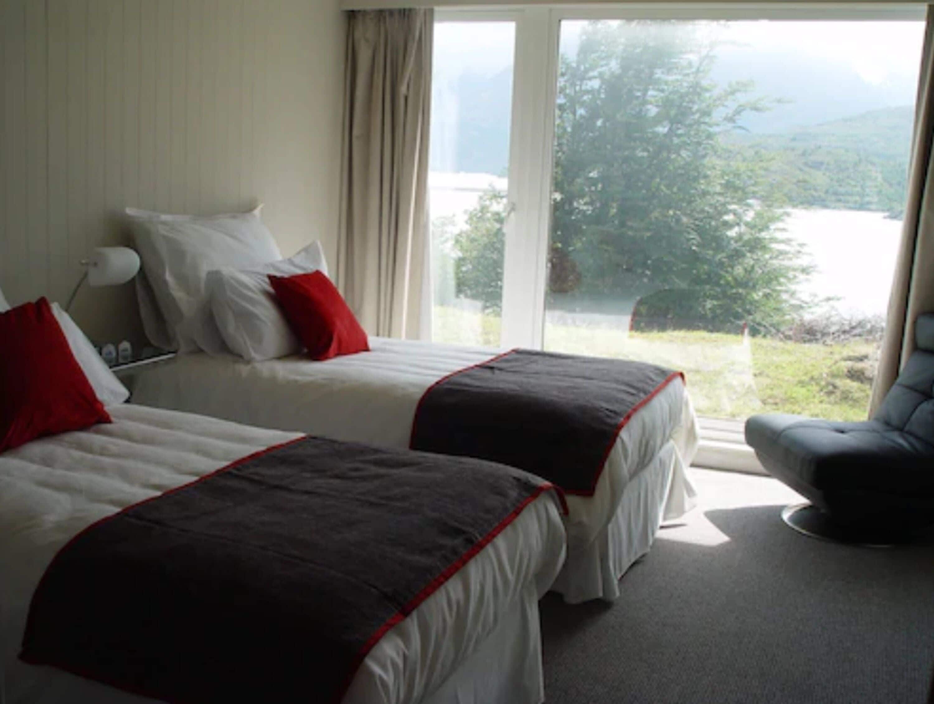 A hotel room with two beds, a chair, and a large window offering a view of a lake and mountains.