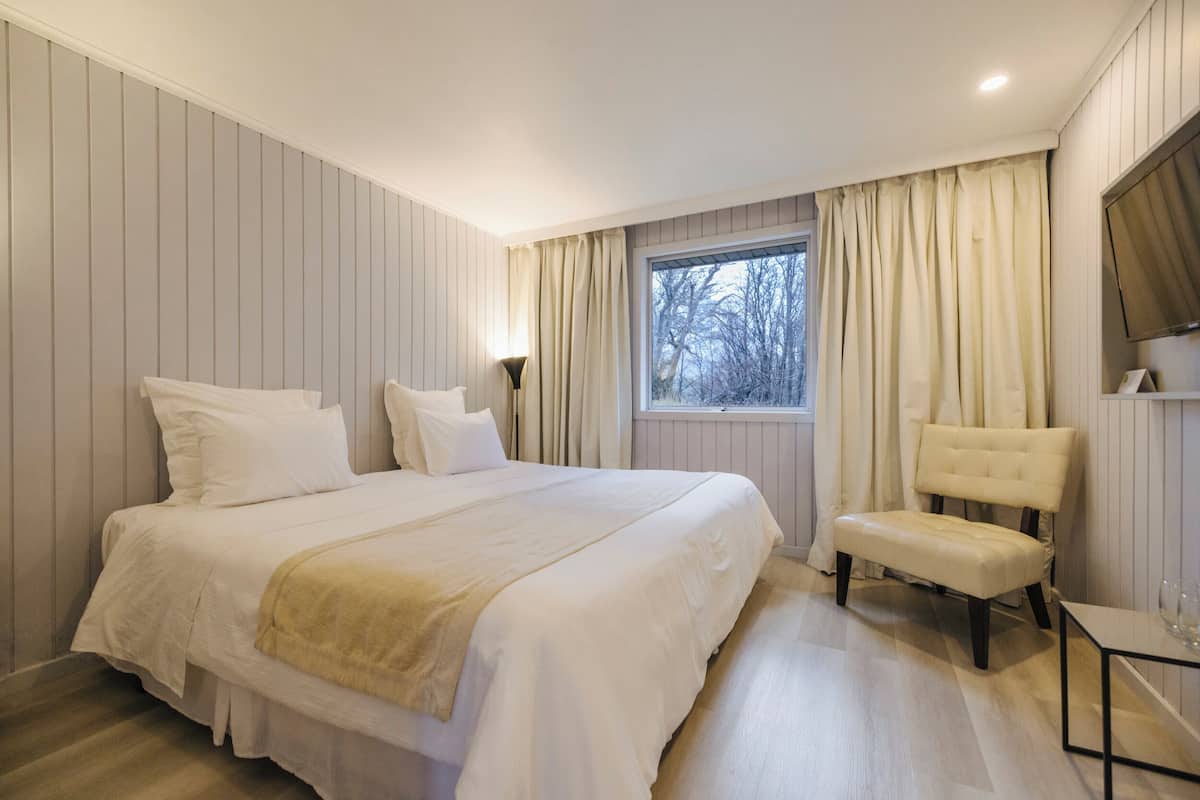 Ein Schlafzimmer mit einem großen Bett, einem Nachttisch, einem Sessel, einem Fenster mit Vorhängen und einem an der Wand befestigten Fernseher.
