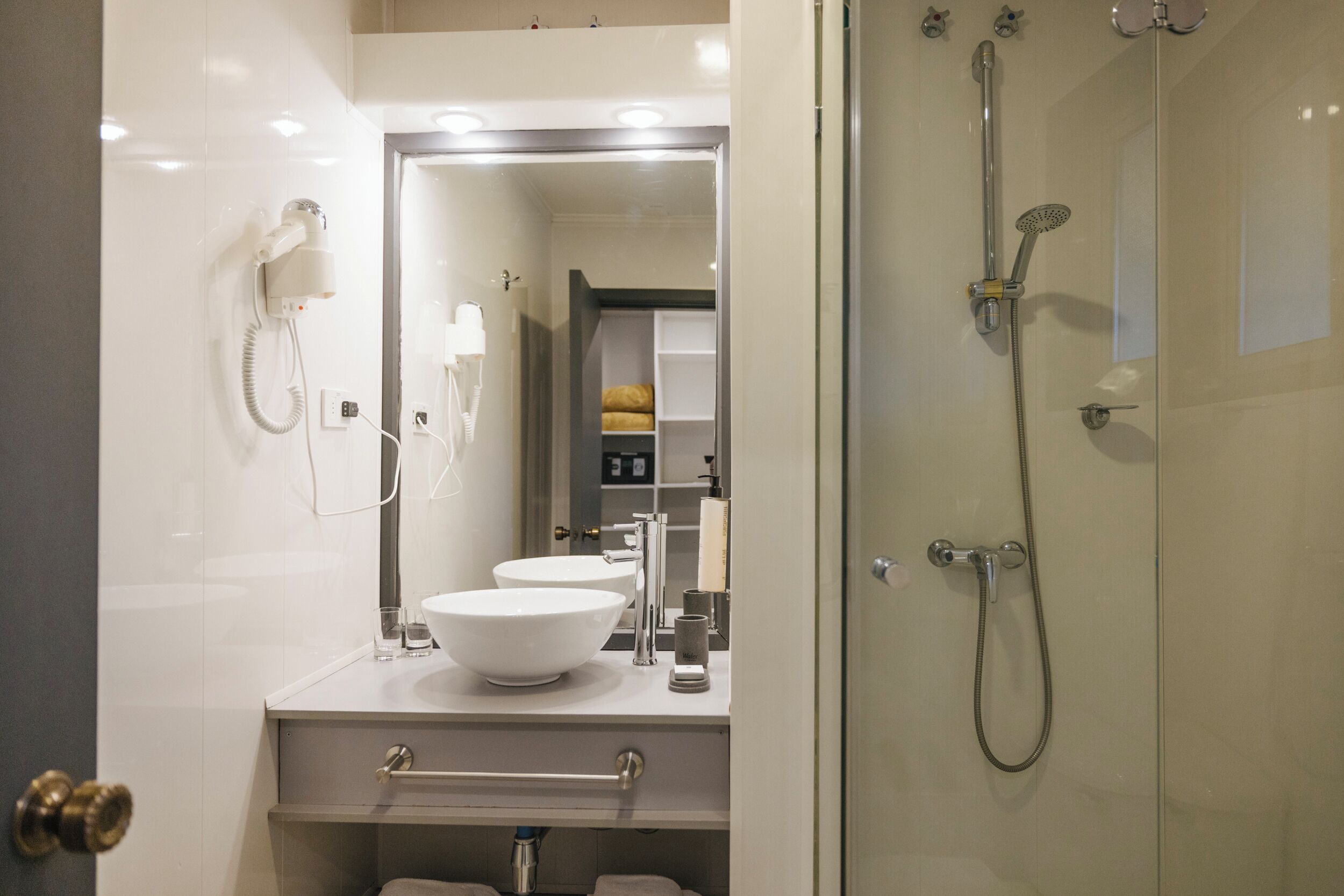 A modern bathroom with a large mirror, a white sink, a shower with a handheld showerhead, and a hairdryer mounted on the wall.